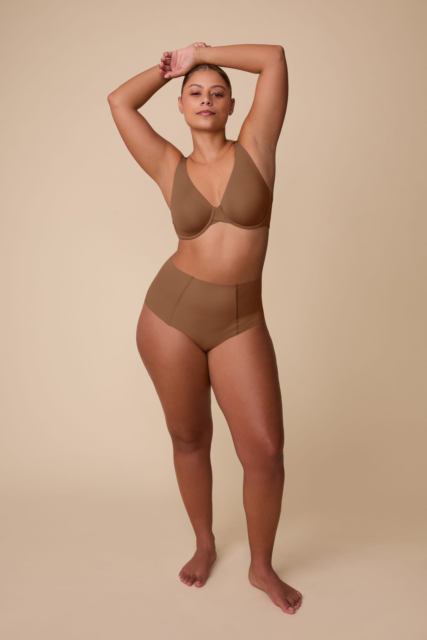 On a neutral beige backdrop, India wears the Base Unlined Bra in Brown, paired with matching underwear, showing the unlined cups, underwire support, and smooth microfiber fabric.