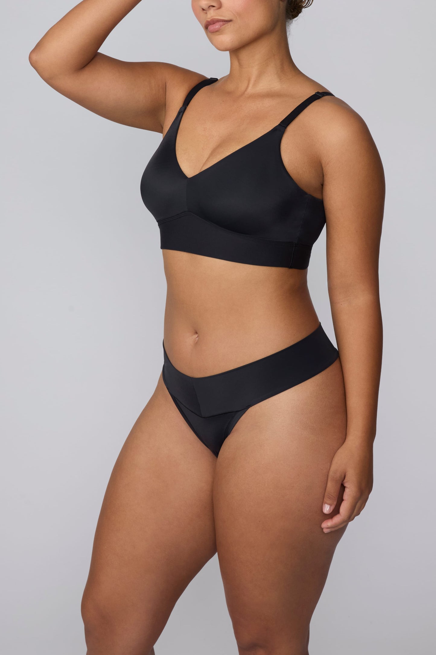 In a studio setting, India wears the Bliss Low Rise Thong in Black with the matching Bliss Bralette in Black. The angled front view highlights the low-rise waistband, smooth fabric, and minimal-coverage thong silhouette.