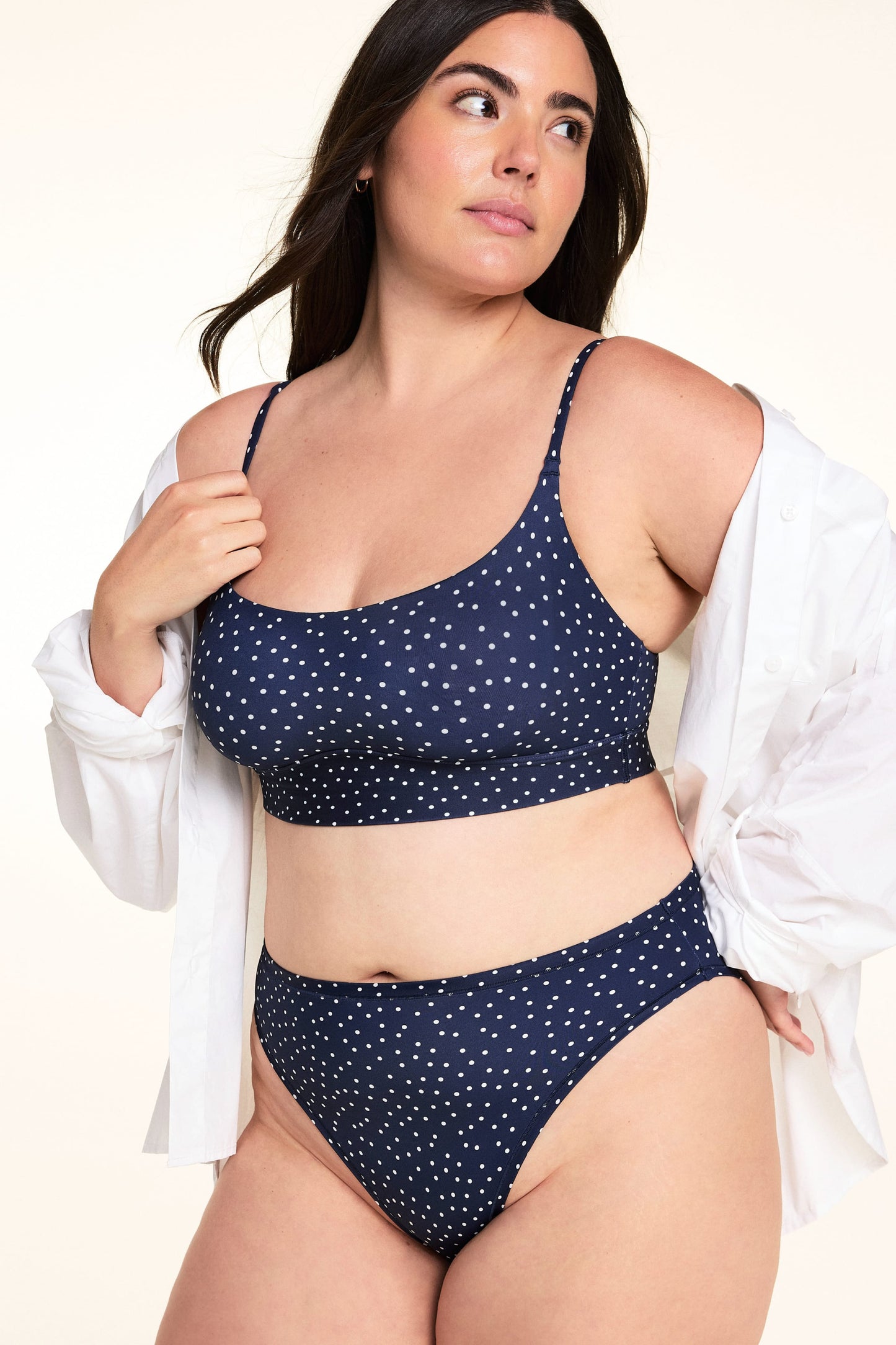 On a light studio backdrop, Alessandra wears the Bliss Scoop Bralette in Navy Dot with matching Navy Dot underwear and an open white button-down shirt draped over her shoulders. The image highlights the scoop neckline, adjustable straps, and soft printed fabric.