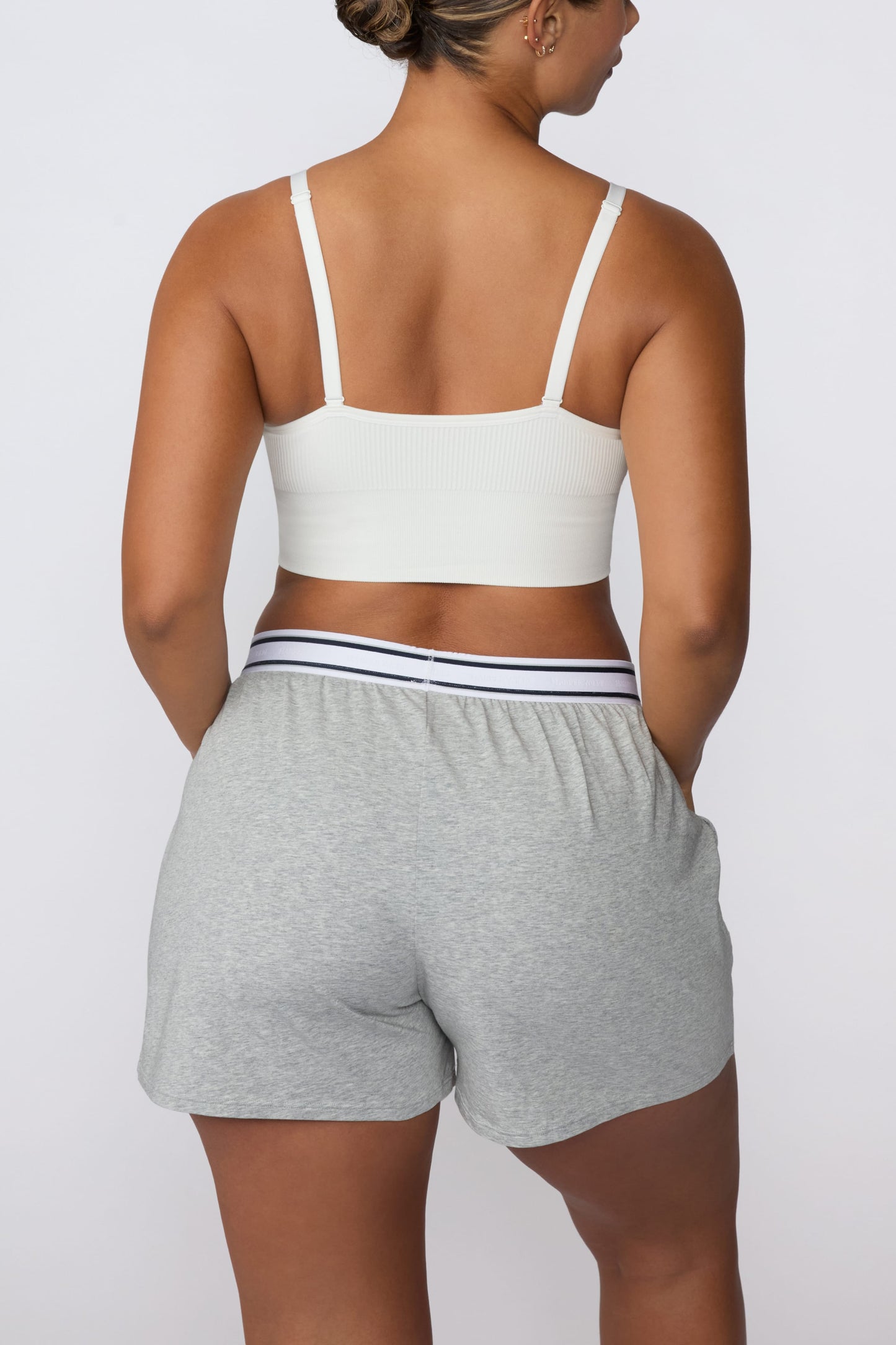 On a light gray studio backdrop, India wears the Cloud Cotton Pajama Short in Heather Grey with a white ribbed bralette. The back view showcases the relaxed silhouette, striped elastic waistband, and soft cotton fabric.
