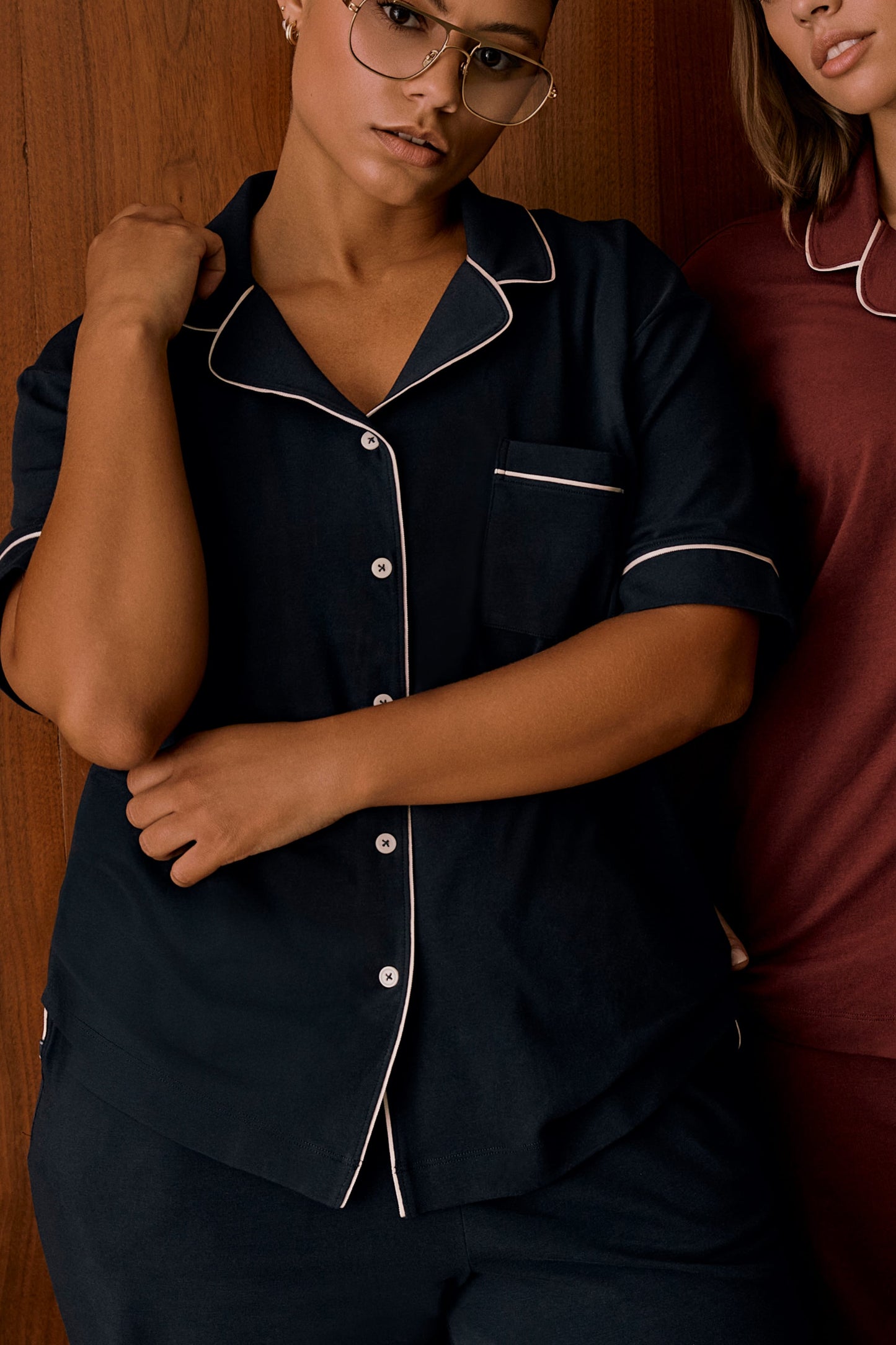 In an indoor setting with a wood-paneled backdrop, India wears the Perfect Pima Pajama Shirt in Navy. The close-up view highlights the button-front closure, chest pocket, contrast piping, and relaxed fit. Another model wearing a burgundy pajama top is partially visible beside her.