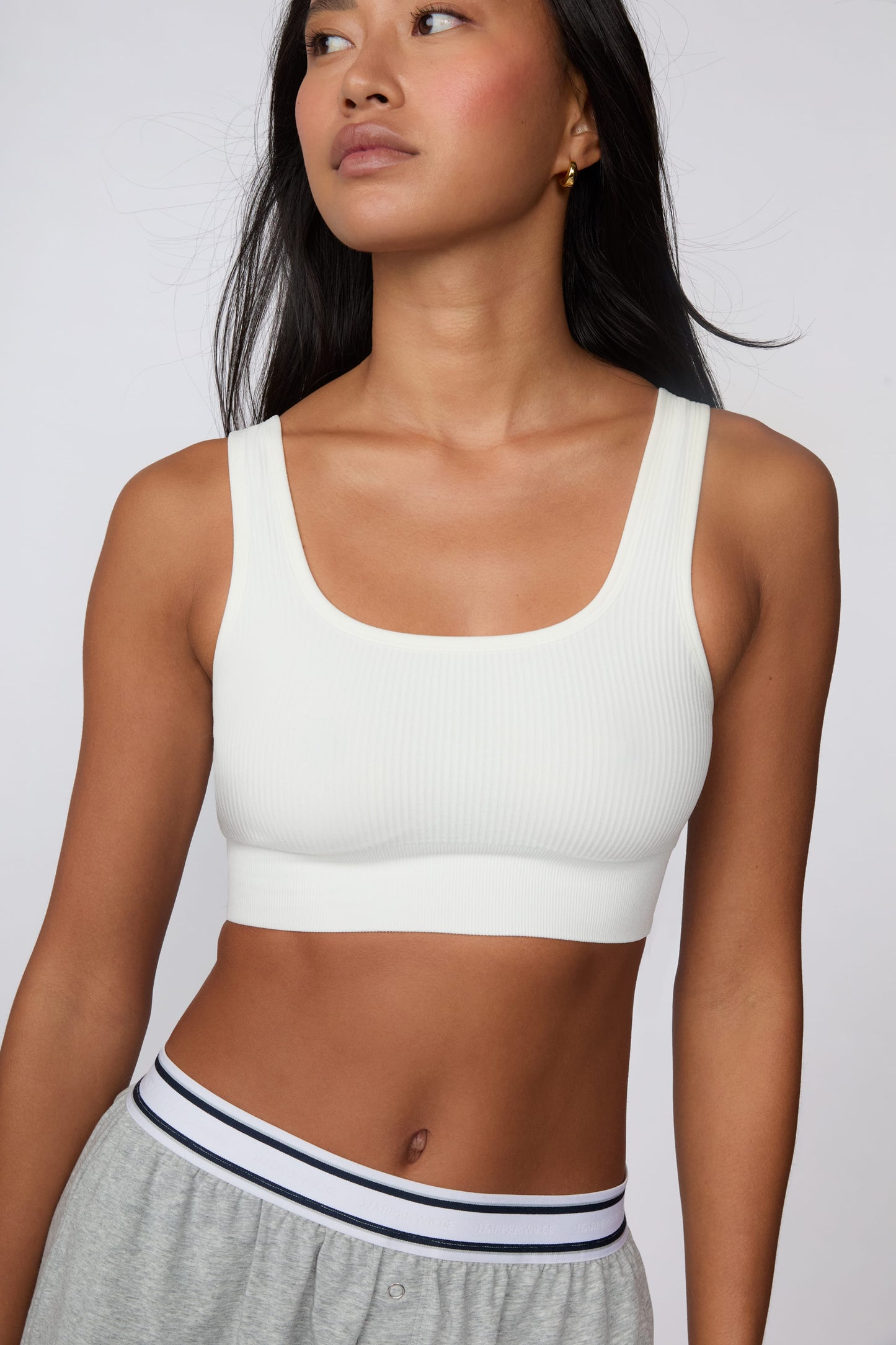 In a studio setting, Amber wears the Ribbed Seamless Scoop Bralette in White with gray lounge shorts. A cropped front view highlights the scoop neckline, wide straps, ribbed texture, and supportive underband.