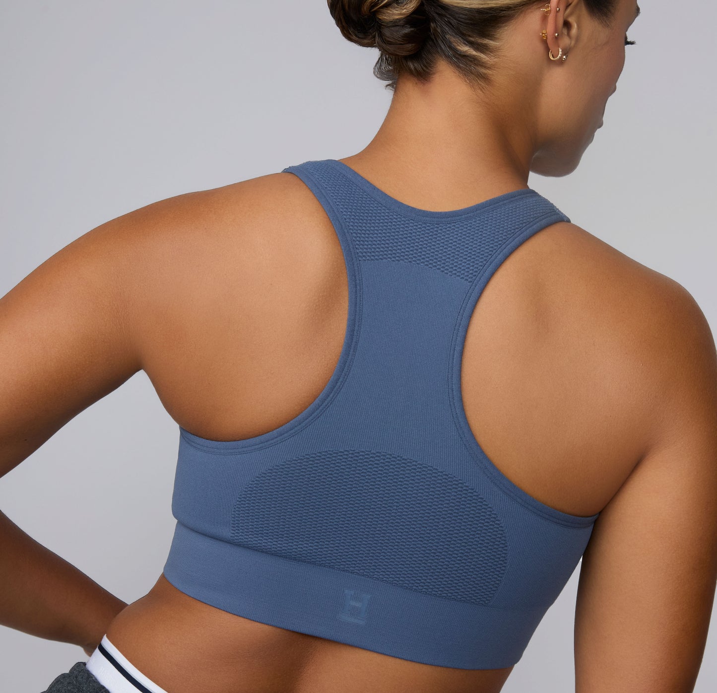 On a light gray studio backdrop, India wears the Seamless Racerback Sports Bra in Evernight, shown from the back to highlight the racerback silhouette, breathable knit-in mesh panels, and wide underband. The smooth, seamless construction is shown for comfortable support during low-impact movement and everyday wear.