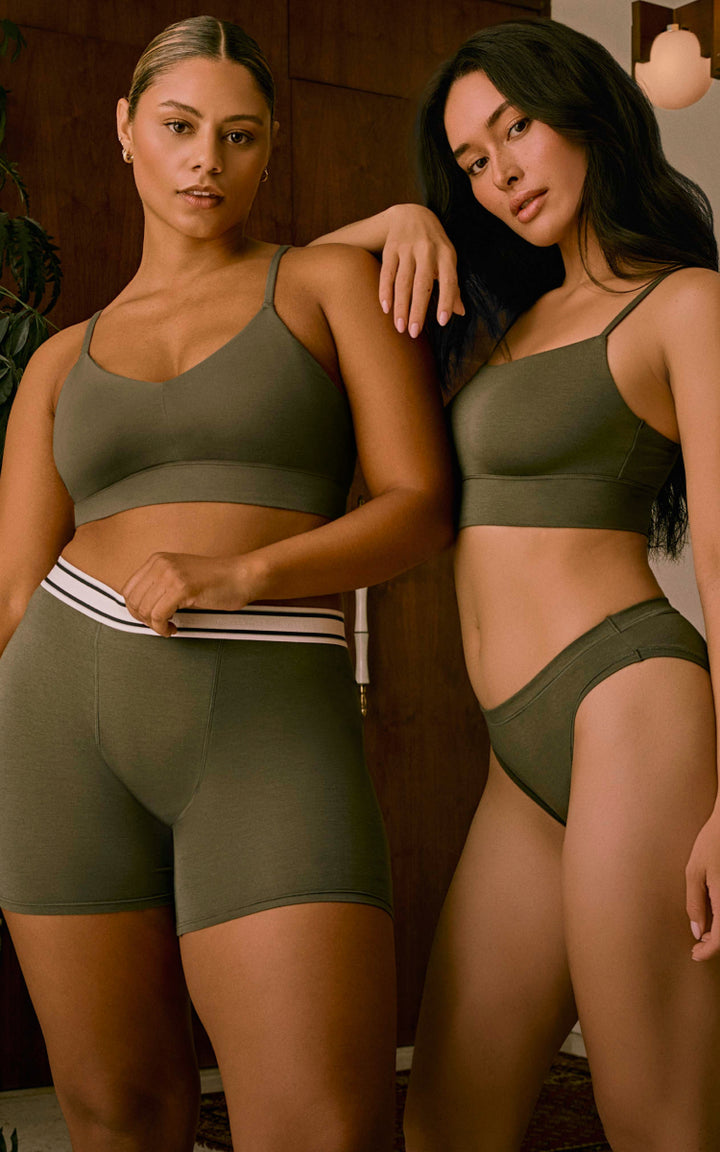 Two models stand side by side indoors, both wearing Cloud Cotton styles in Olive. The model on the right wears the Thin Strap Bralette and matching Bikini. The model on the left wears the Cloud Cotton Triangle Bralette and Logo Boxer Brief, featuring a striped waistband and longer inseam for full coverage comfort.