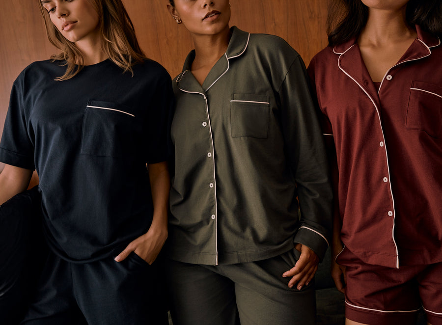 Three models wear Harper Wilde Pima Pajamas in Navy, Olive, and Bordeaux. The model on the left wears a Navy short sleeve tee and pants set, the center model wears a full button-down Olive set, and the model on the right wears a Bordeaux short sleeve button-down and matching bralette underneath. All garments feature signature white piping and soft, breathable Pima cotton fabric.