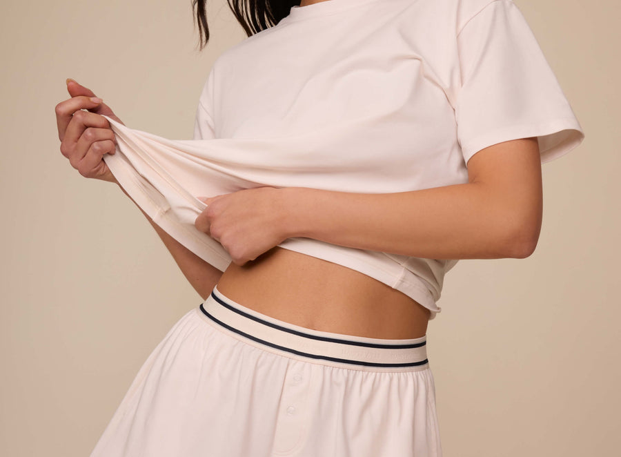 Close-up of a model lifting the hem of a black Cloud Cotton Sleep T-shirt to reveal the coordinating Quartz sleep shorts. The waistband is elastic with Harper Wilde branding and dark contrast stripes, showcasing the soft stretch and casual comfort of the fabric.