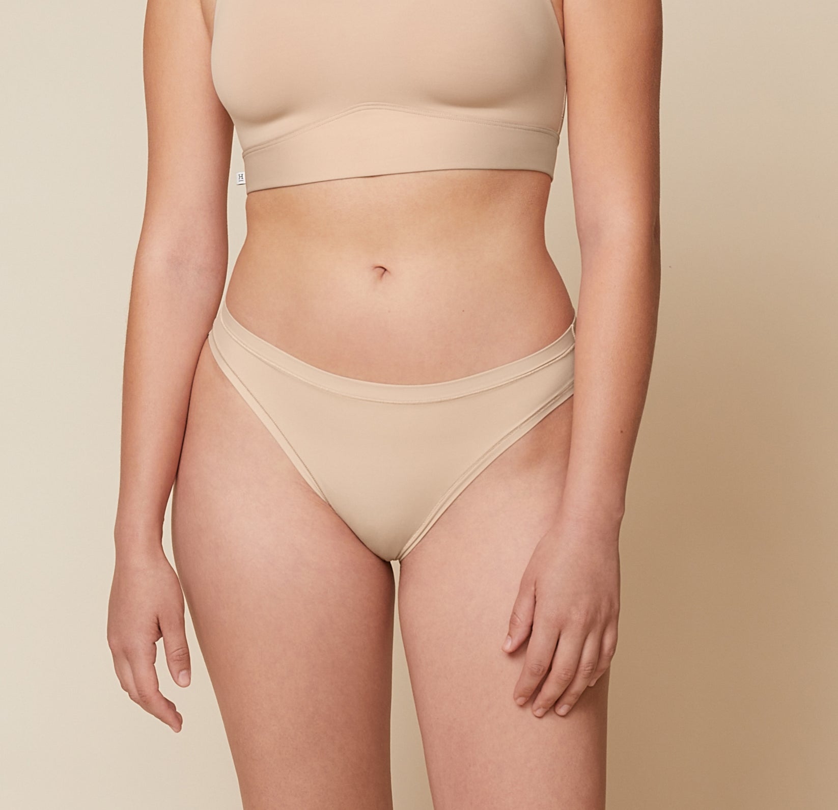 A close-up side view of Carli, focusing on her lower body. She is wearing the Bliss Bikini in Beige, showcasing the design and fit of the underwear. sizing: Carli wears size Medium