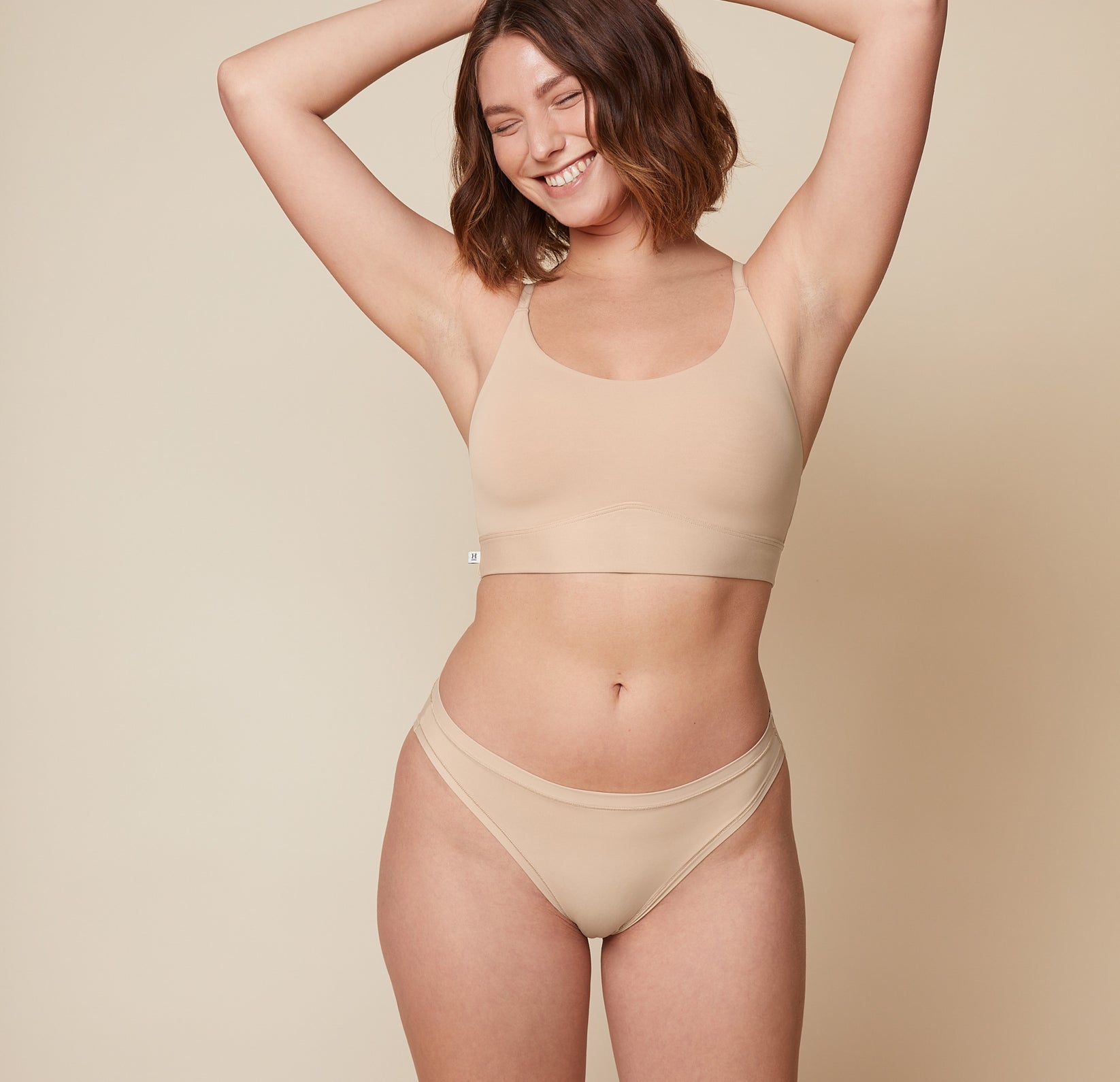 Carli is standing facing forward with her arms raised above her head, smiling. She is wearing the Bliss Racerback Bralette and the Bliss Bikini in Beige, showcasing the set from the front. sizing: Carli wears size Medium
