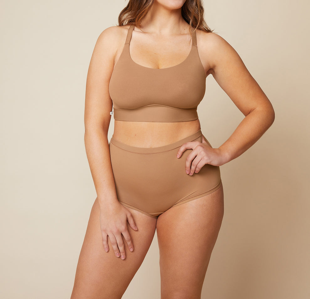 Model standing with her left hand on her hip and her right arm down by her side. She’s wearing the Bliss Racerback Bralette and matching Bliss Boyshort in the color Tan. sizing: Summerlyn wears size Large