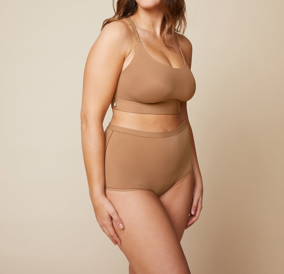 Model standing with her arms down by her side. She’s wearing the Bliss Racerback Bralette and matching Bliss Boyshort in the color Tan.