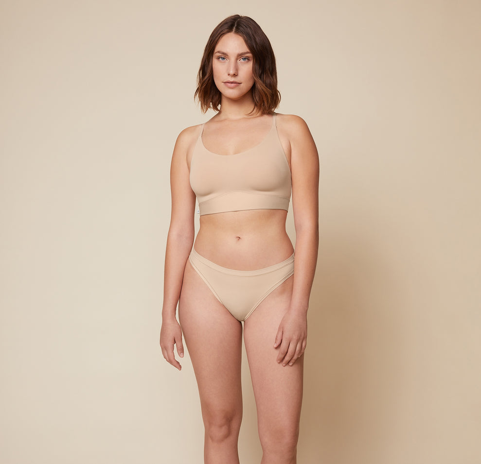 Carli is standing facing forward with her hands resting by her sides. She is wearing the Bliss Racerback Bralette and the Bliss Bikini in Beige, providing a full front view of the set.