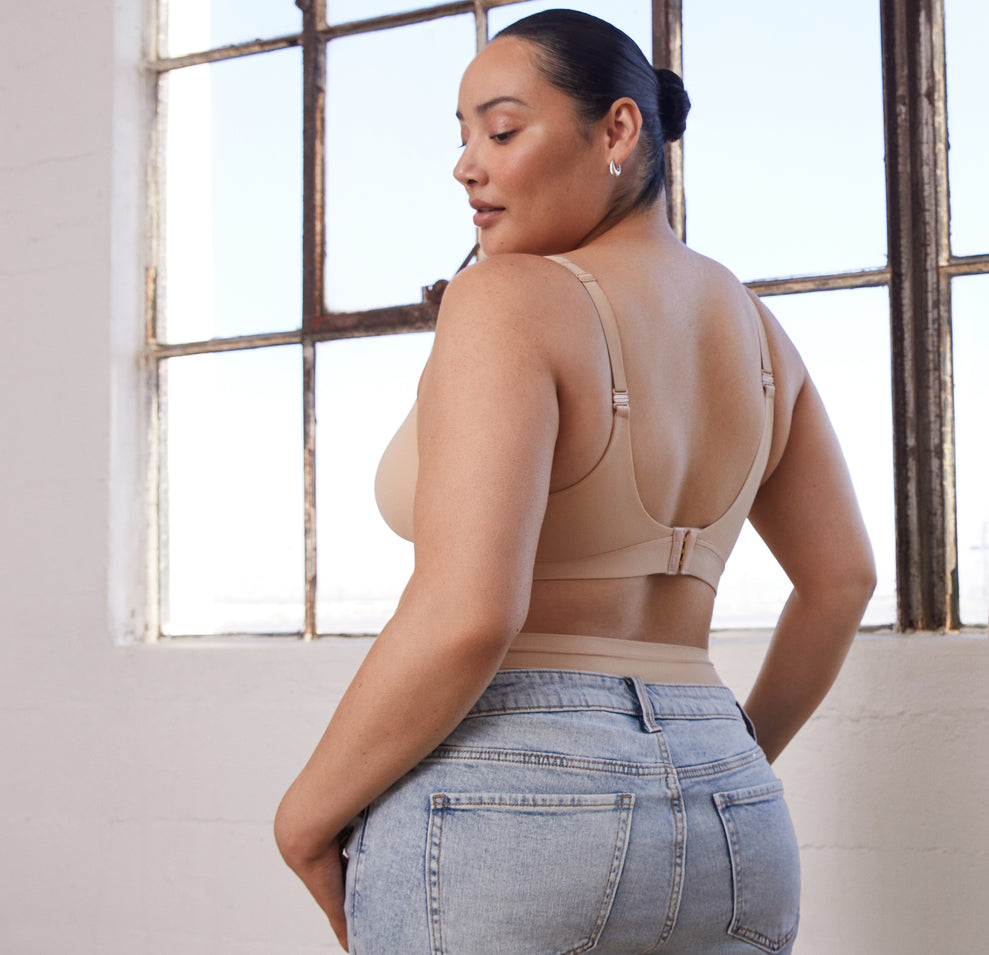 Lifestyle image of Bianca in a warehouse, wearing the Bliss Plunge Adjustable Bralette and Bliss Highwaist Brief in Beige, with casual jeans, showing how the intimates can be styled for every day.
