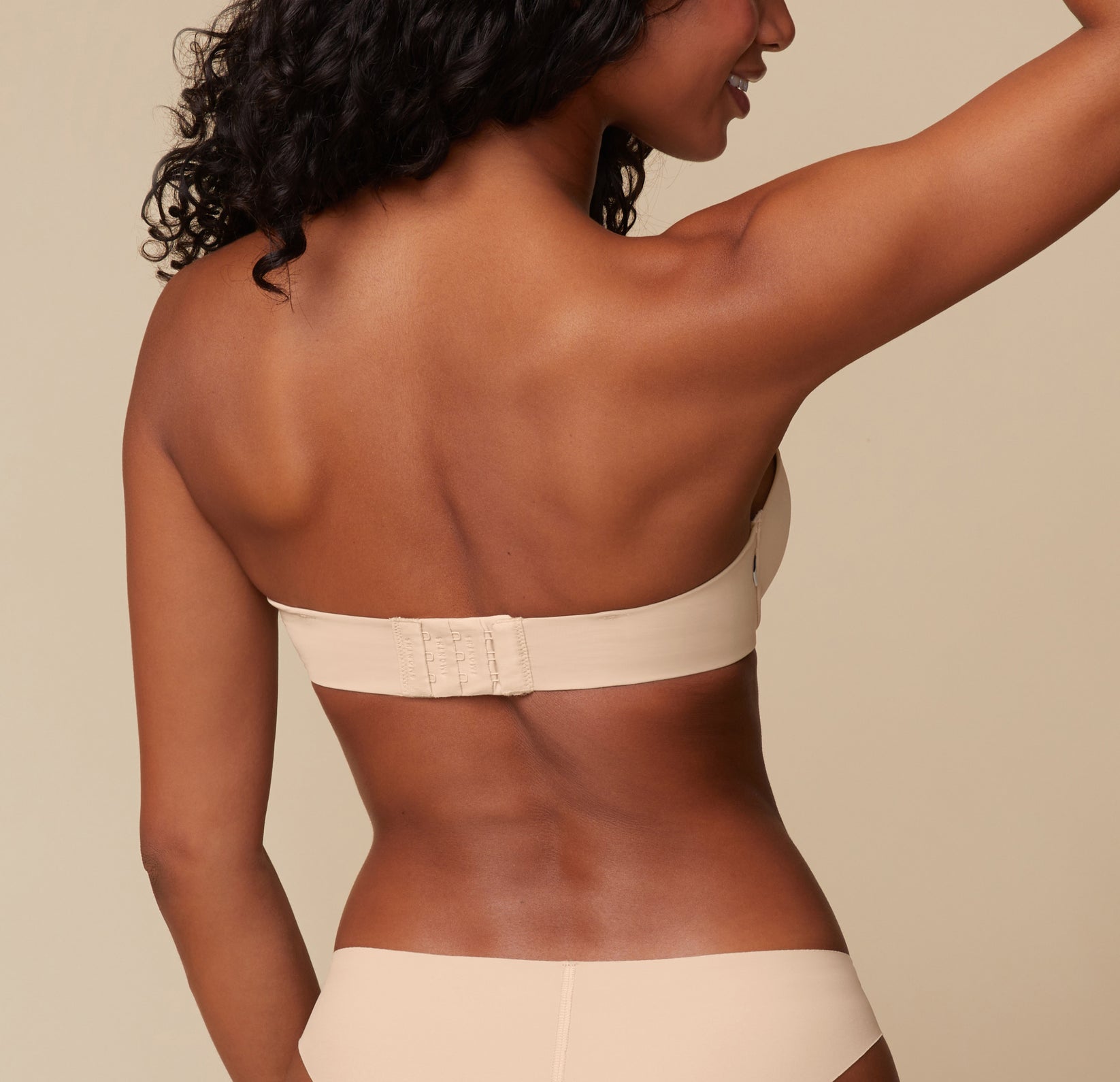 Back view of Ally modeling the Base Strapless Bra in Beige. She poses with her arms lifted, emphasizing the back closure and smooth band of the strapless bra.