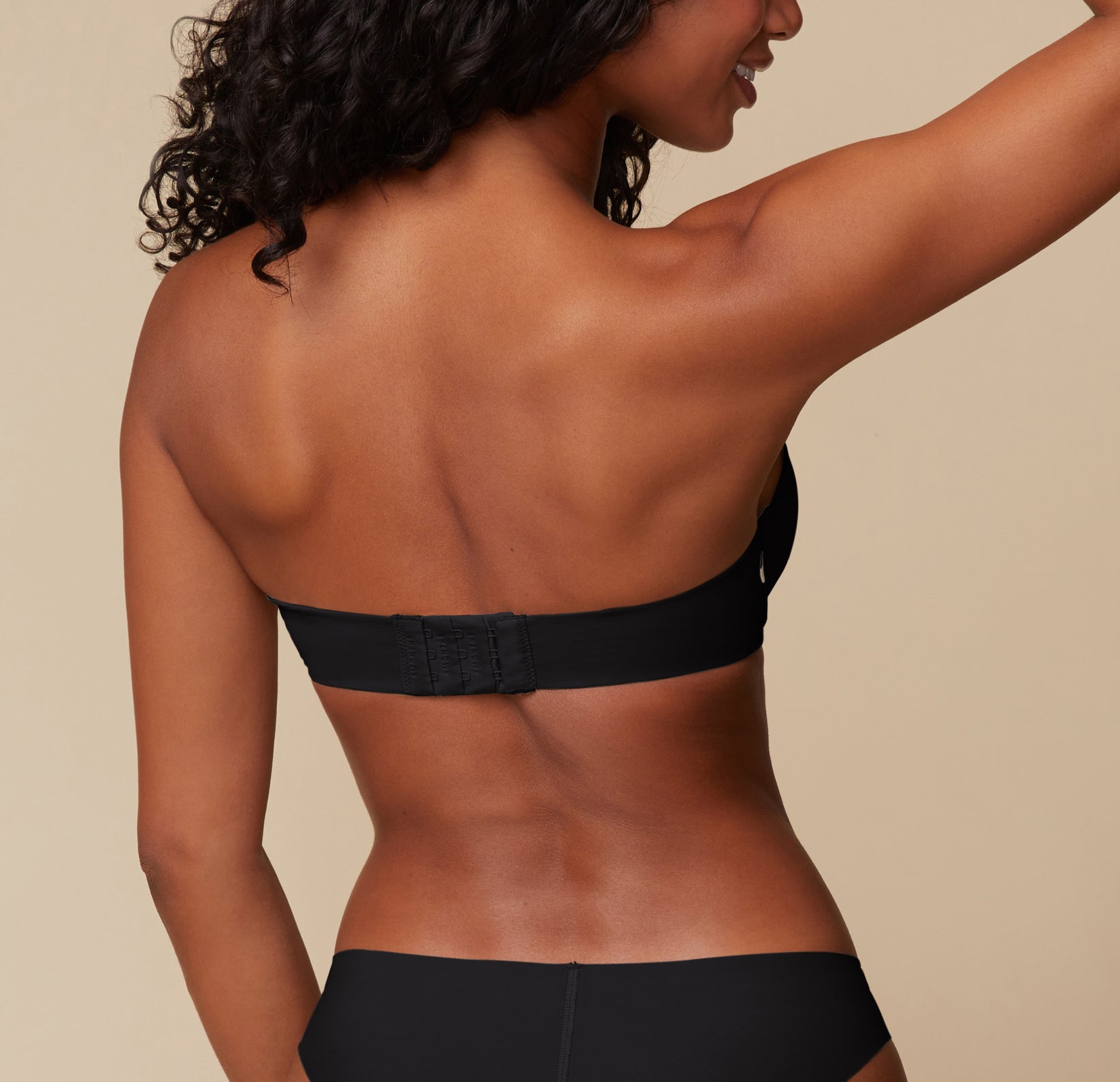 Back view of Ally modeling the Base Strapless Bra in Black. She poses with her arms lifted, emphasizing the back closure and smooth band of the strapless bra.
