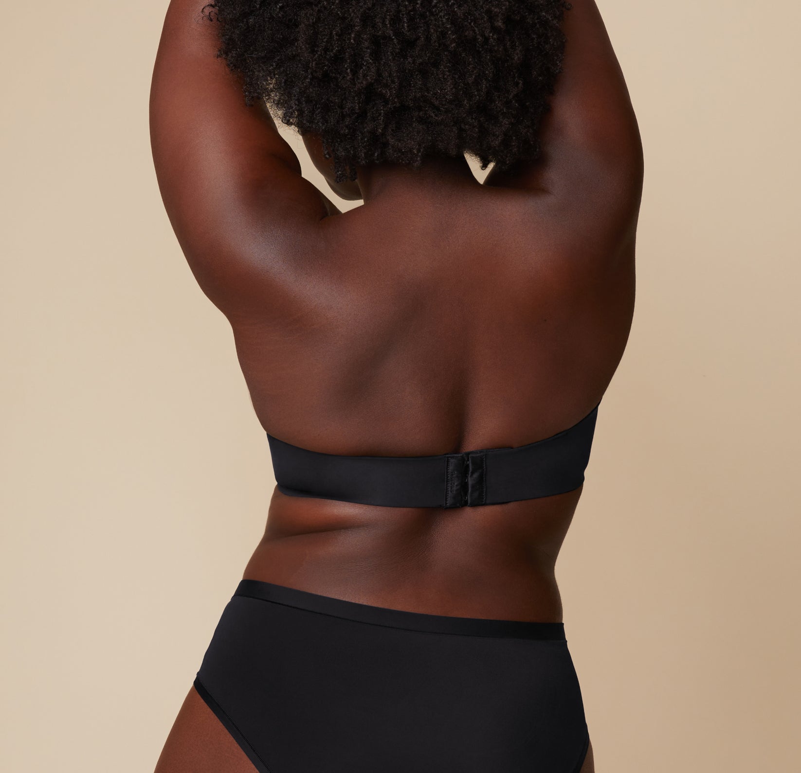 Back view of Akesha modeling the Base Strapless Bra in Black. Her arms are lifted, showing the strapless bra's back closure and band.