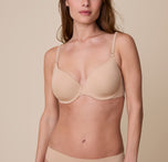 Gracie wears the Base T-Shirt Bra in Beige, facing forward with a soft expression. The image highlights the smooth, lightly lined cups, underwire support, and adjustable straps against a neutral beige background. sizing: Gracie wears size 32D