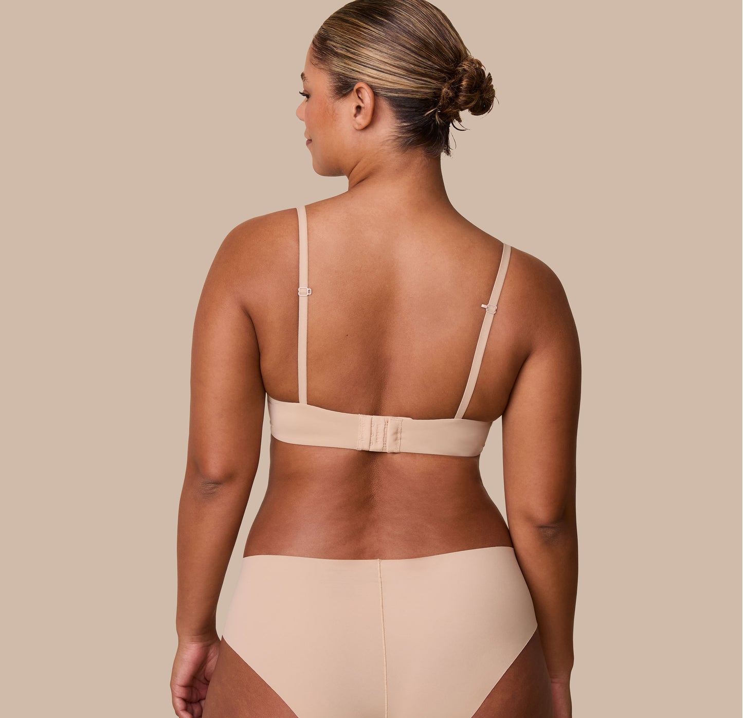India faces away from the camera, displaying the back of the Base T-Shirt Bra in Beige with its wide band, hook-and-eye closure, and convertible racerback feature.
