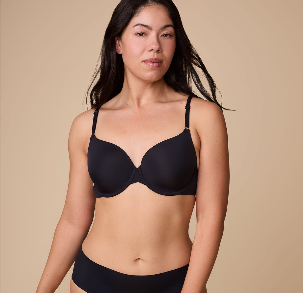 Emersyn models the Base T-Shirt Bra in Black, standing in a relaxed pose to highlight the smooth, form-fitting design. sizing: Emersyn wears size 36C