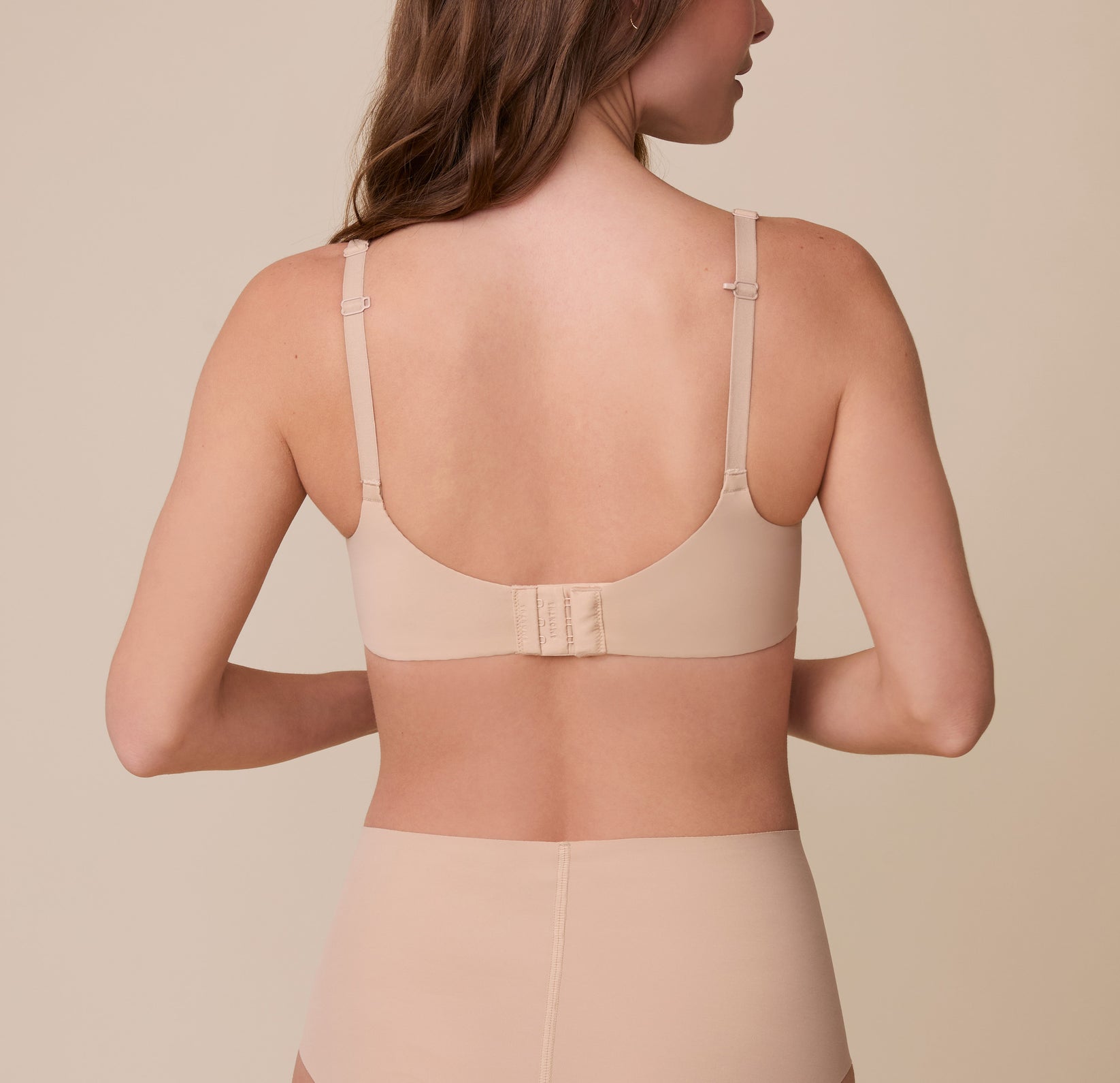 Gracie showcases the back of the Base Unlined Bra in Beige, revealing the wide band, hook-and-eye closure, and convertible racerback feature for a supportive, customizable fit.