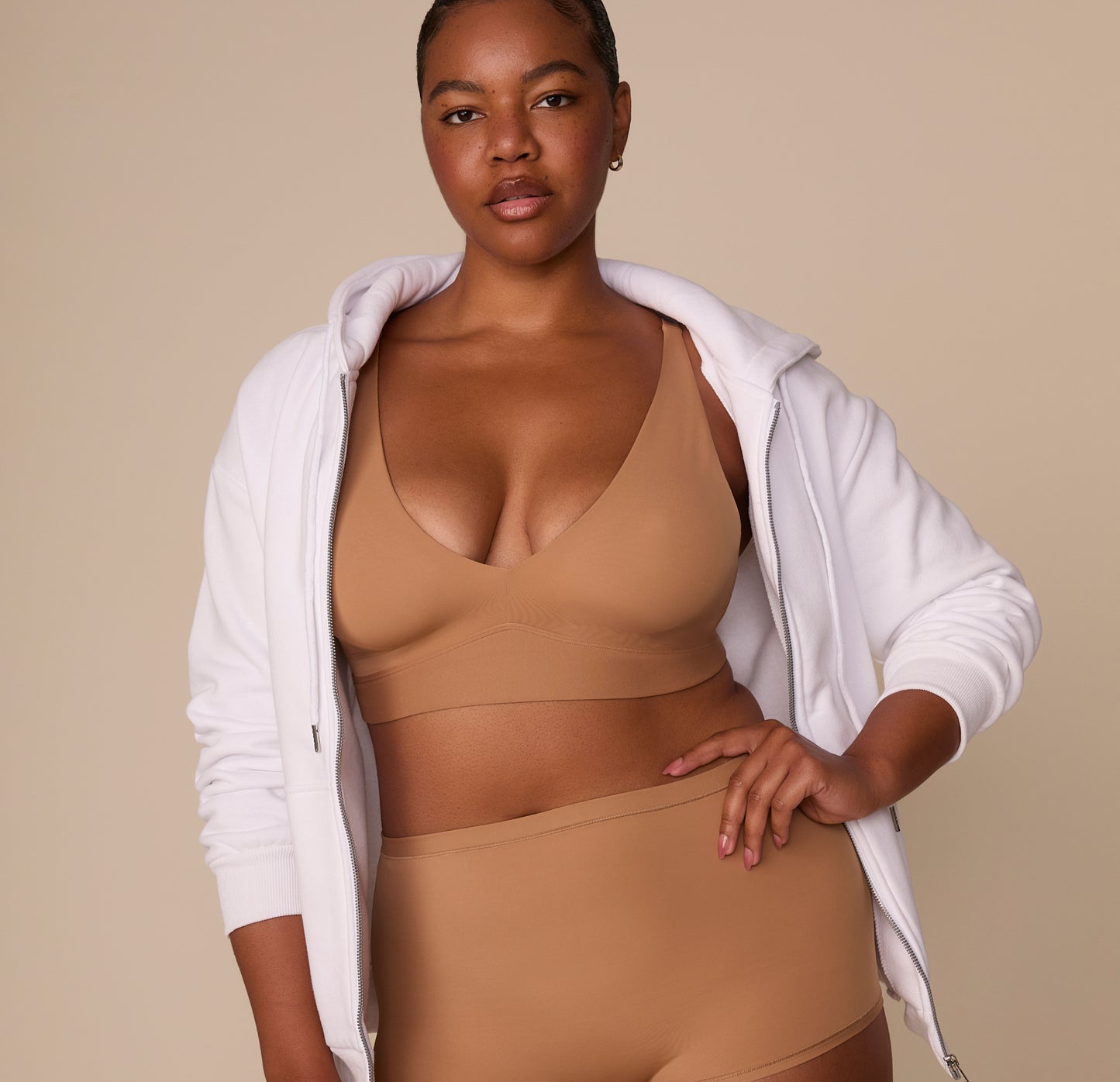 Zarria styled in the Bliss Plunge Bralette in Tan paired with high-rise bottoms and a white zip-up hoodie worn off-shoulder.
