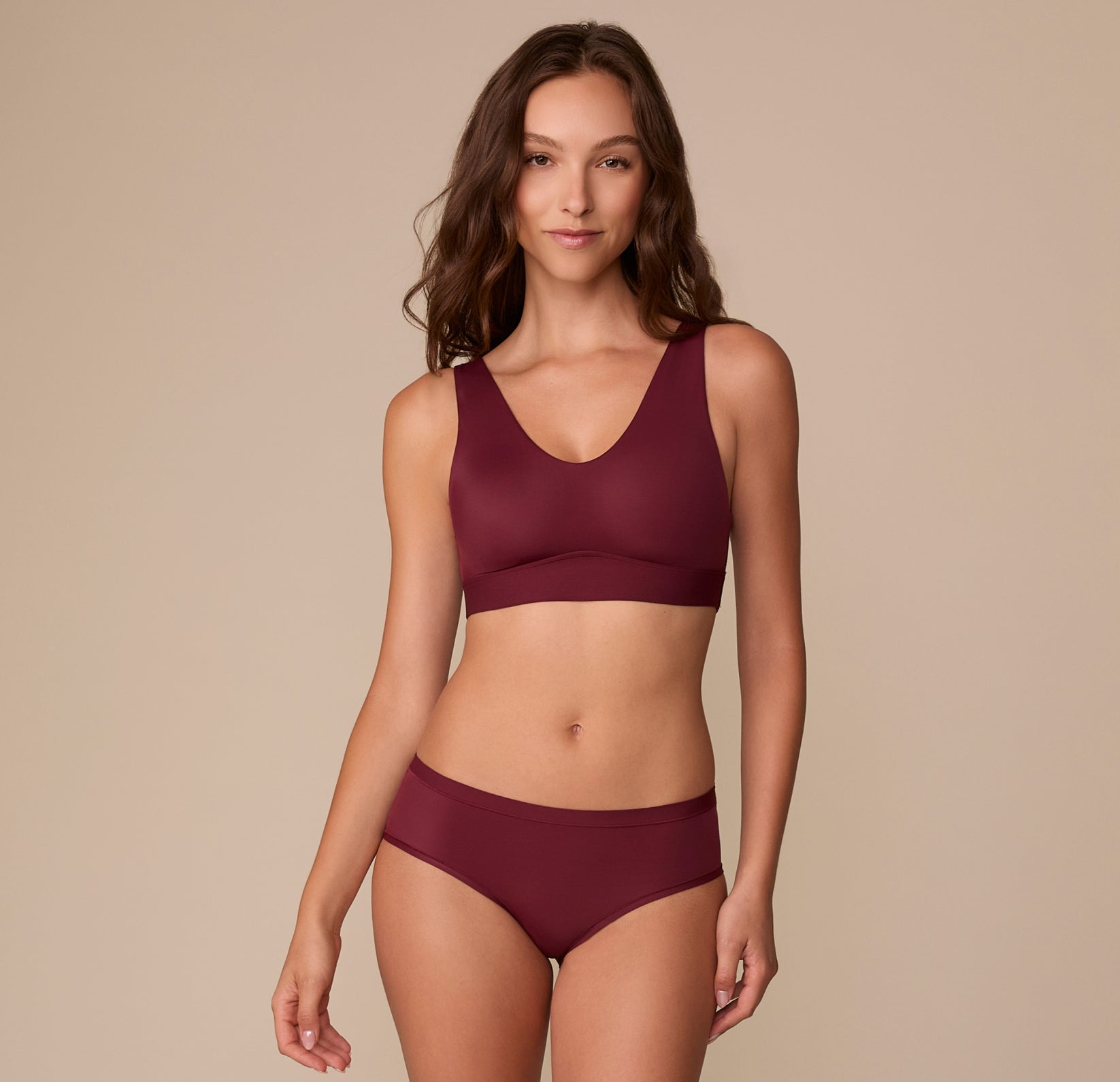 Full body view of Alexis modeling the Bliss Bralette and coordinating Bliss Hiphugger in Bordeaux against a beige background.