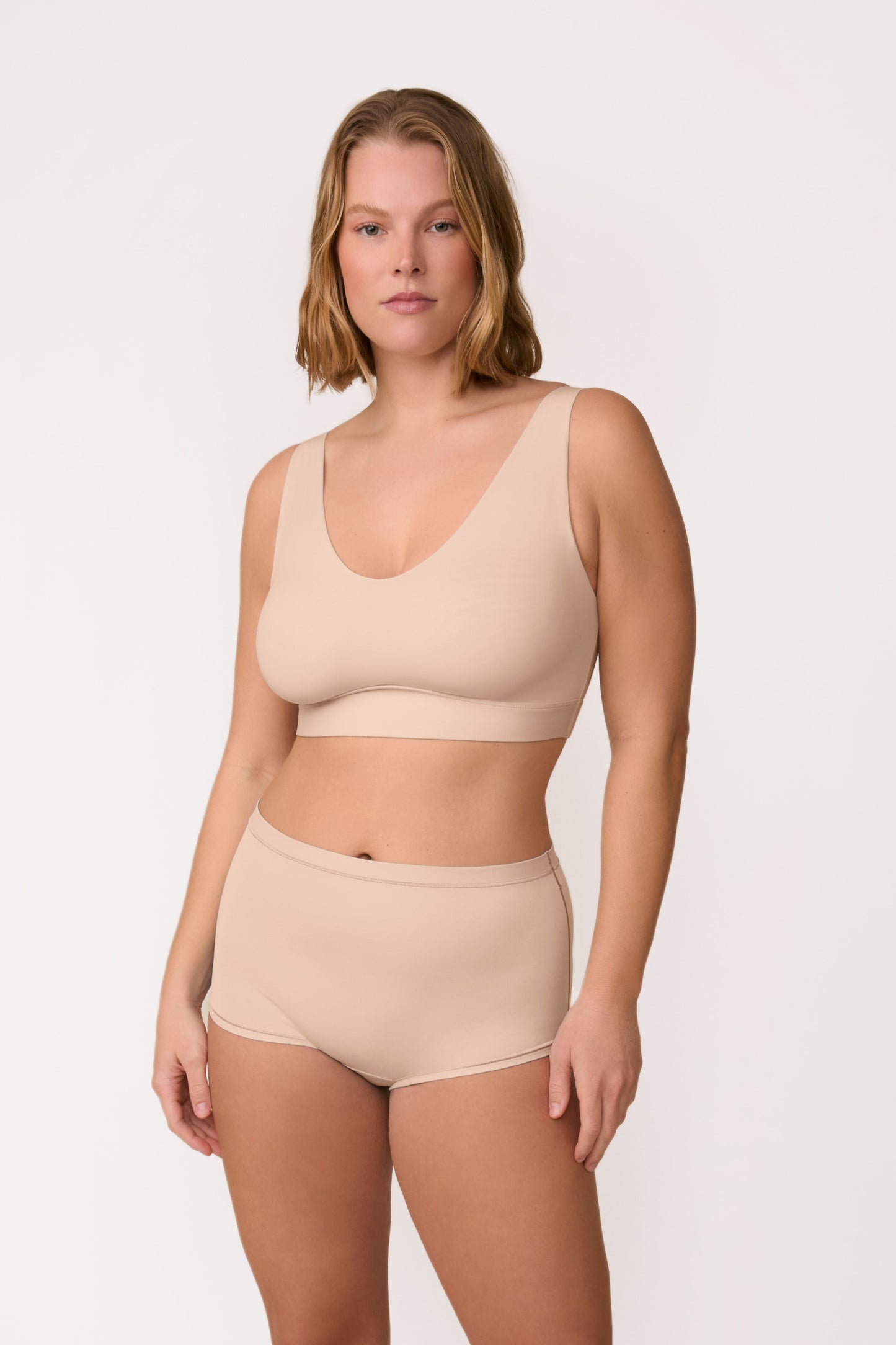On a light neutral studio backdrop, Sonny wears the Bliss Bralette in Beige with matching underwear, shown in a full-body view. The pullover bralette has a scoop neckline and wireless band designed for soft, all-day support.