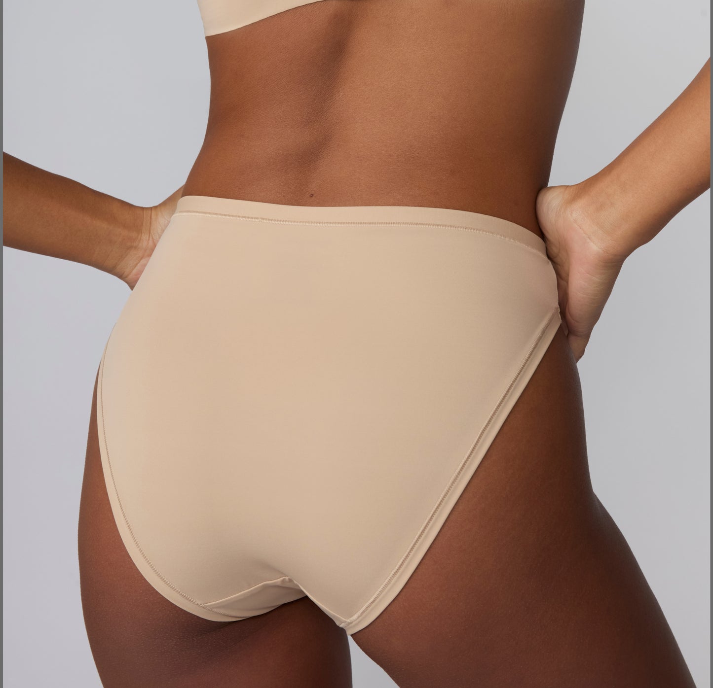 On a light gray studio backdrop, a back view of Amber wearing the Bliss French Cut Brief in Beige shows the smooth rear coverage and seamless-looking edges under the fabric.