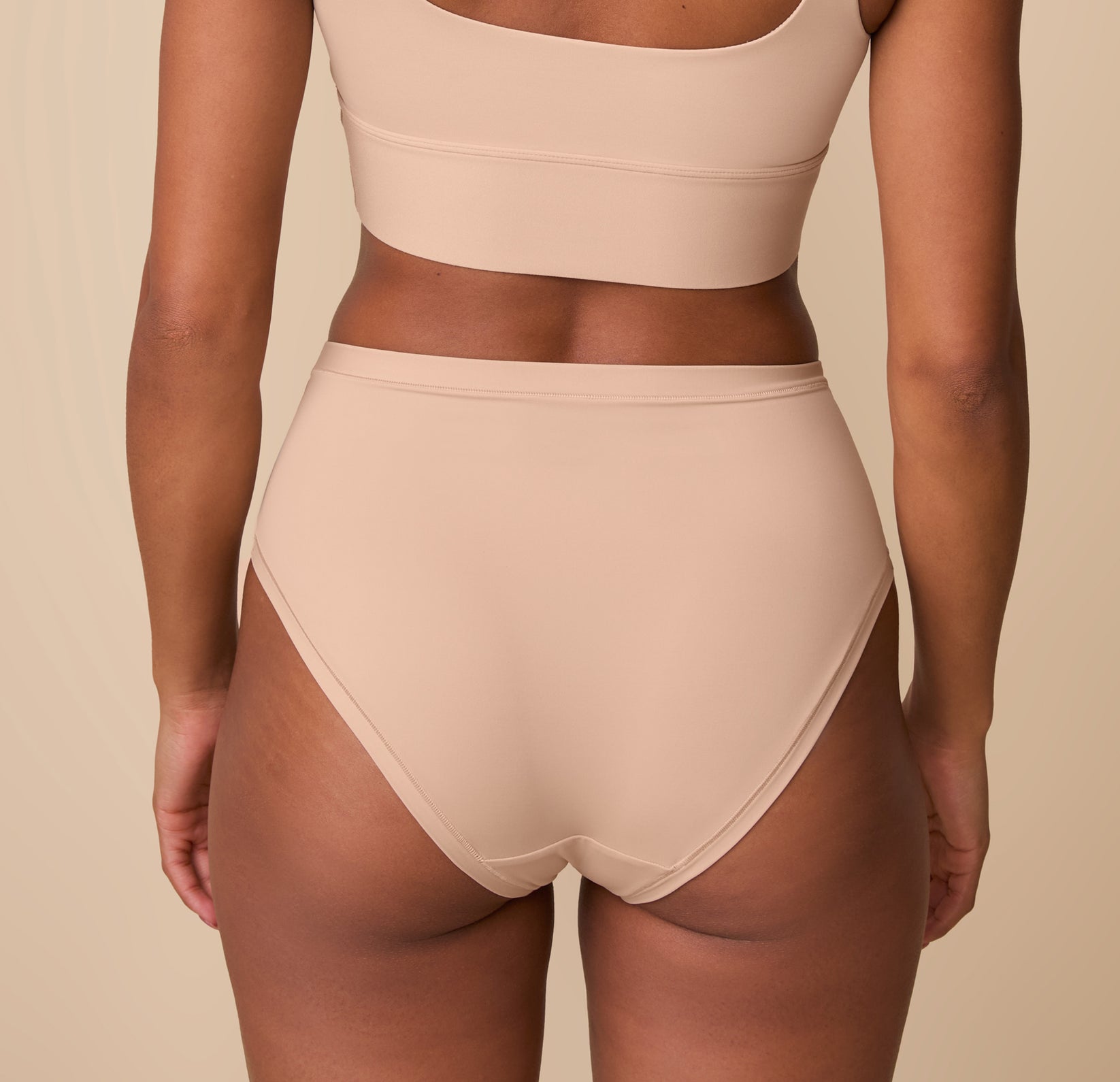 Back view of Ally wearing the Bliss Highwaist Brief in Beige, standing against a soft beige background. The brief features a high-rise fit with smooth, minimal edges for all-day comfort and a flattering silhouette.