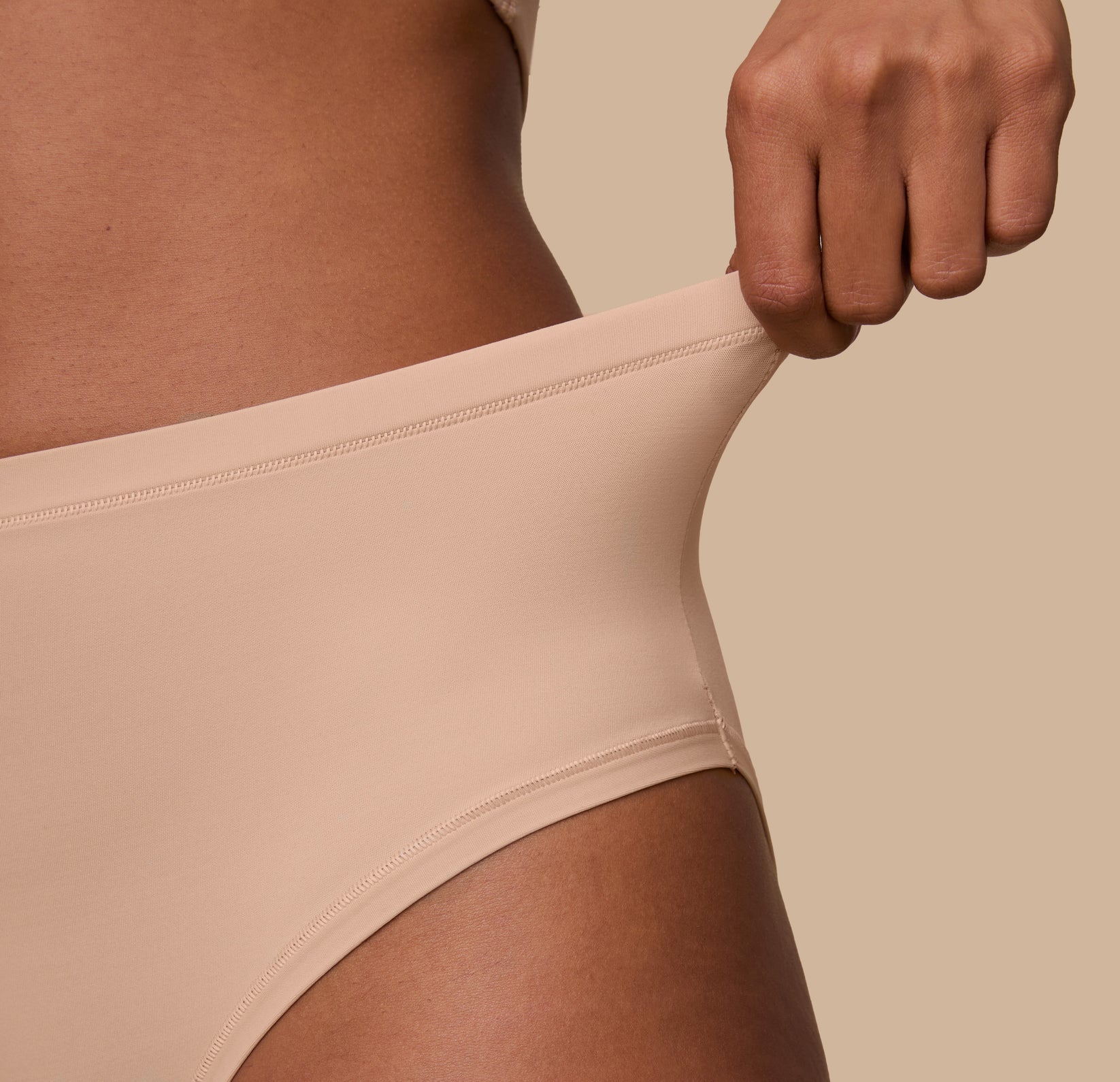 A close-up of Ally gently pulling the waistband of the Bliss Highwaist Brief in Beige to demonstrate the flexible, stretch-fit fabric that moves with the body.