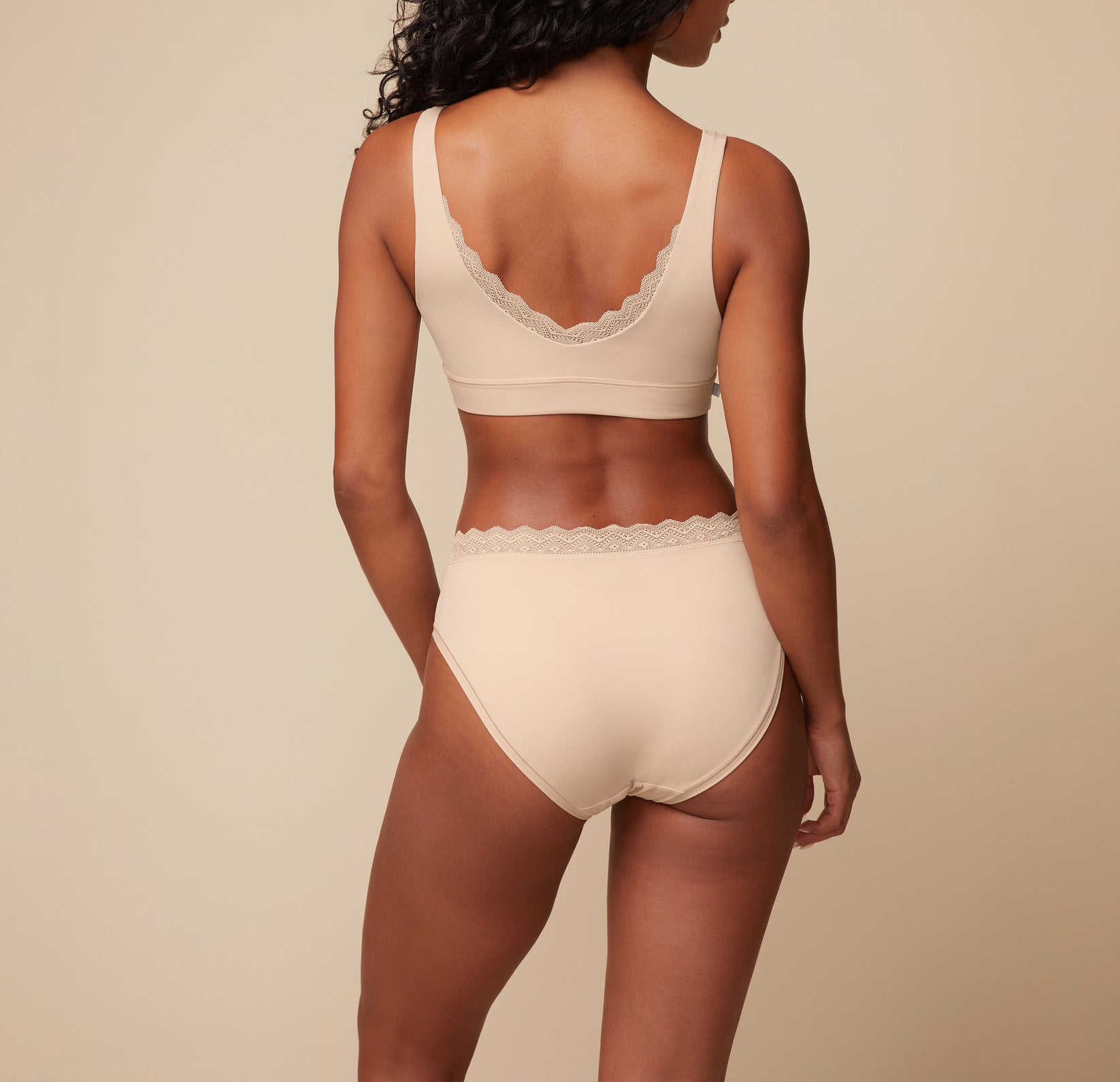 Back view of Ally wearing the Bliss Lace Highwaist Brief and matching Bliss Lace Bralette in Beige. The image emphasizes the full-coverage design of the brief and the supportive, lace-back design of the bralette.