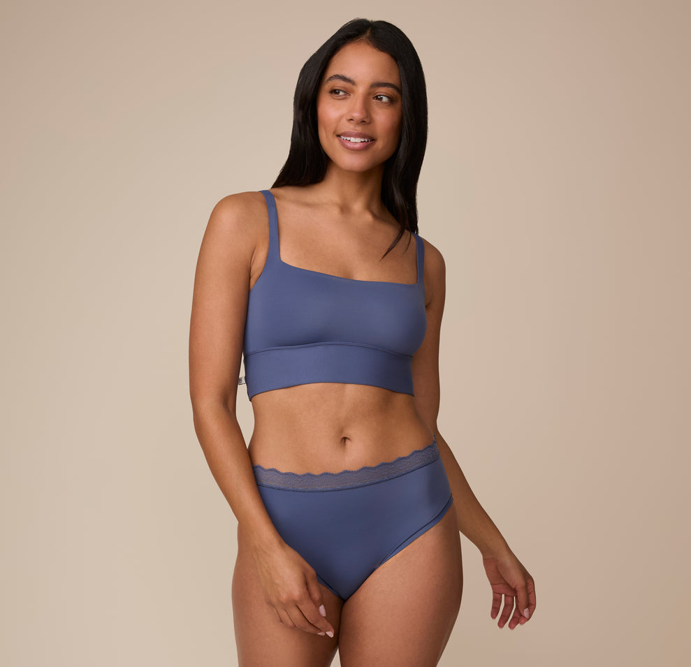Nemesis in a full-body shot wearing the Bliss Lace Highwaist Brief and the Bliss Square Neck Bralette in Twilight. The image showcases the matching set’s sleek, minimal seam fabric and tonal lace accents.
