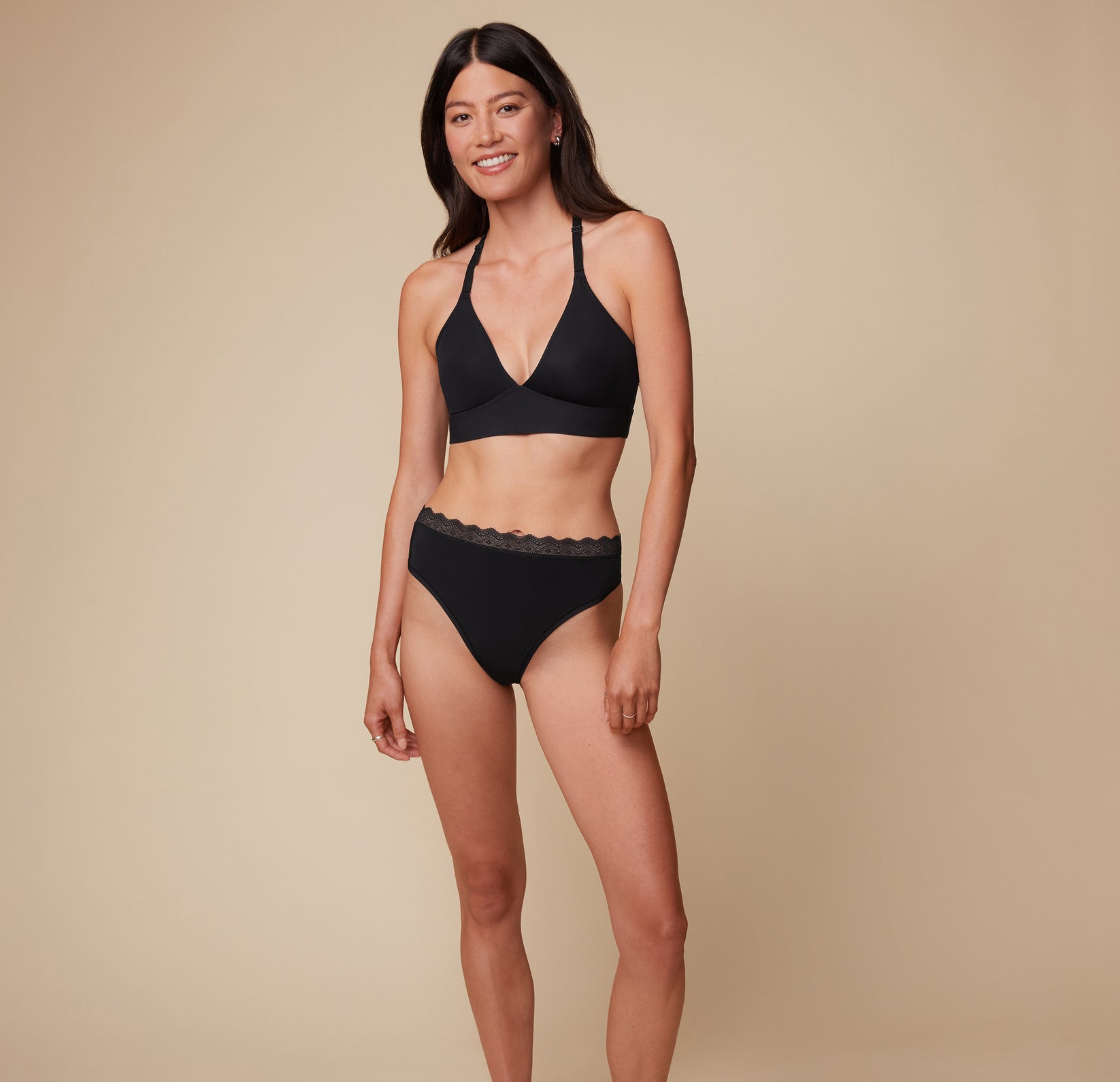 Model Anna wearing the Bliss Lace Thong in Black, paired with the matching Bliss Lace Halterneck Bralette. She stands facing forward, highlighting the minimal coverage fit of the lace thong and the supportive halterneck bralette.