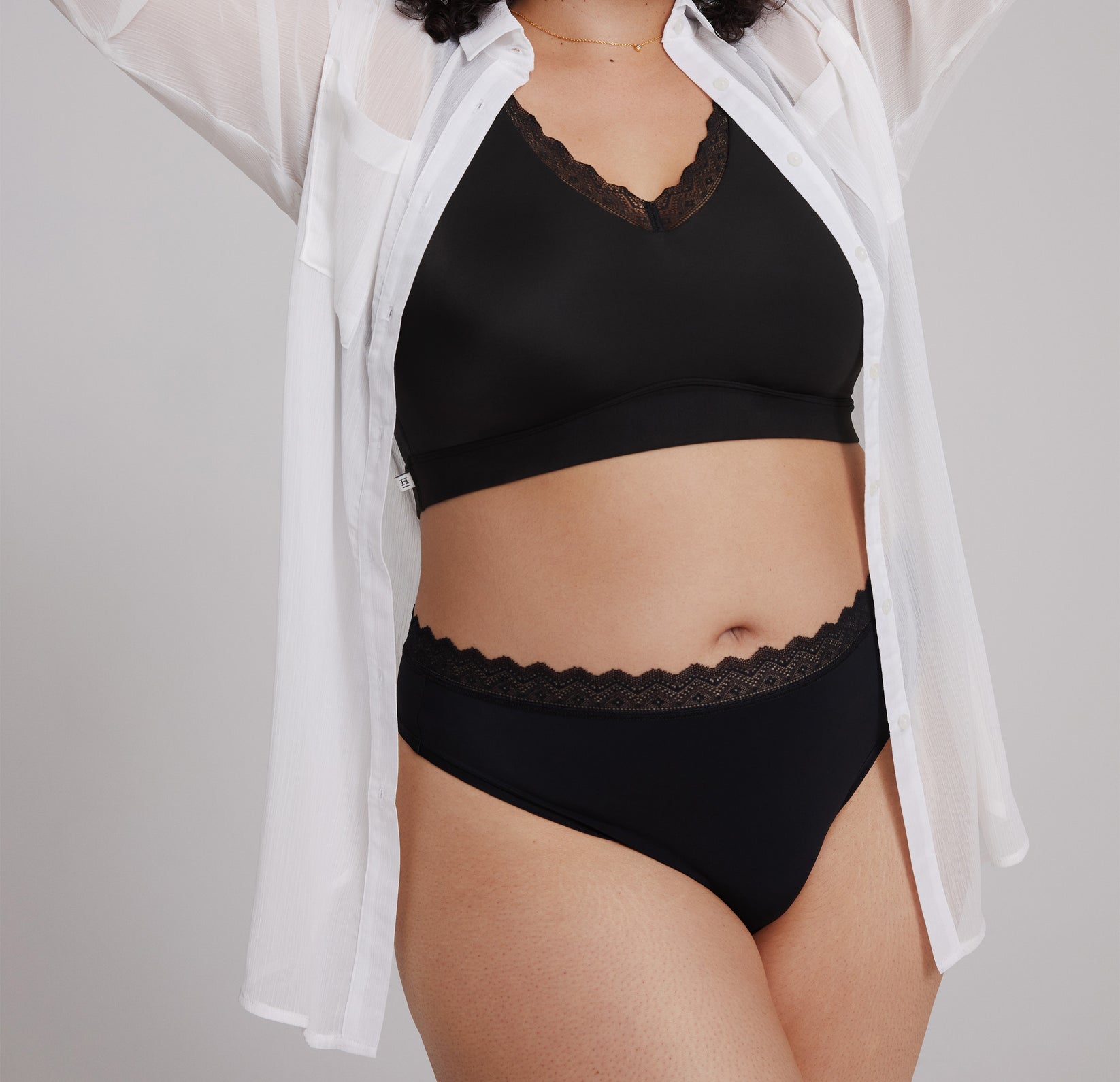 Bianca wearing the Bliss Lace Thong in Black, styled with a sheer white shirt. The image emphasizes the thong’s comfortable fit and versatile, everyday styling.