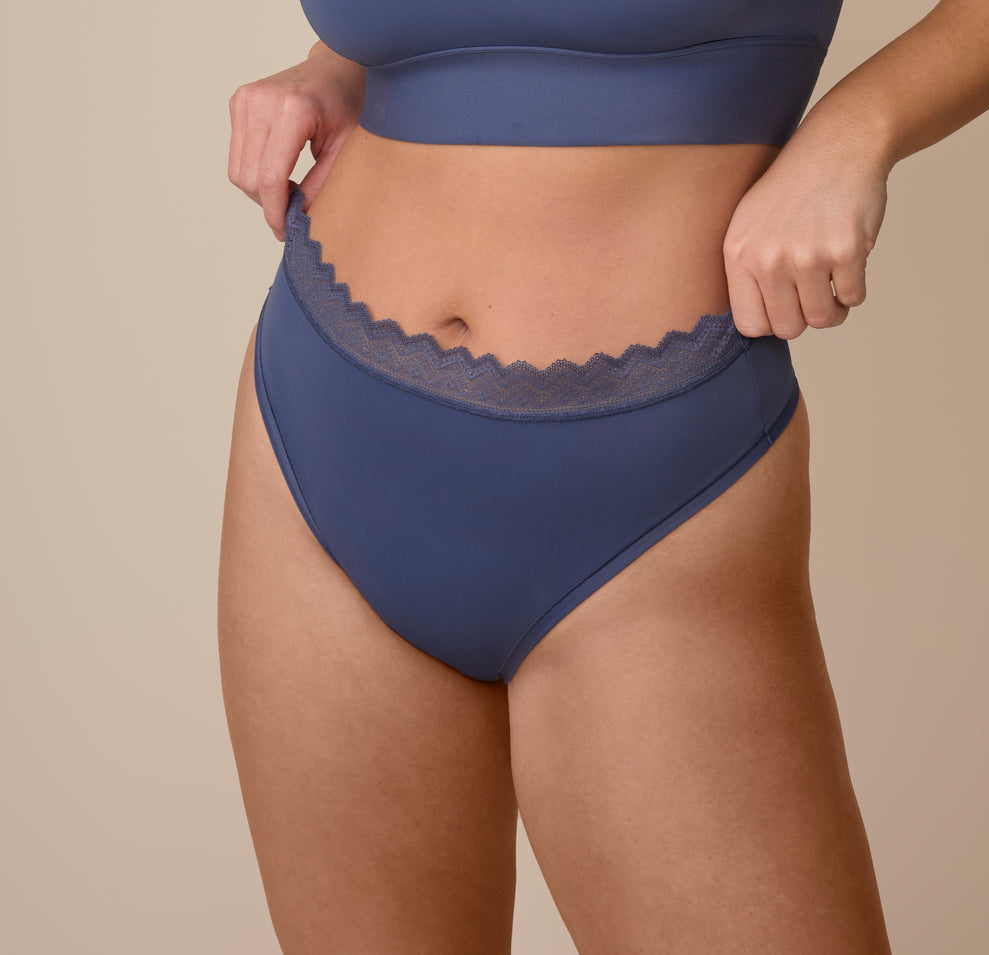 Sonnie adjusting the waistband of the Bliss Lace Highwaist Thong in Twilight. The high-rise waistband is finished with scalloped lace that lays flat without digging in. sizing: Sonnie wears size XL