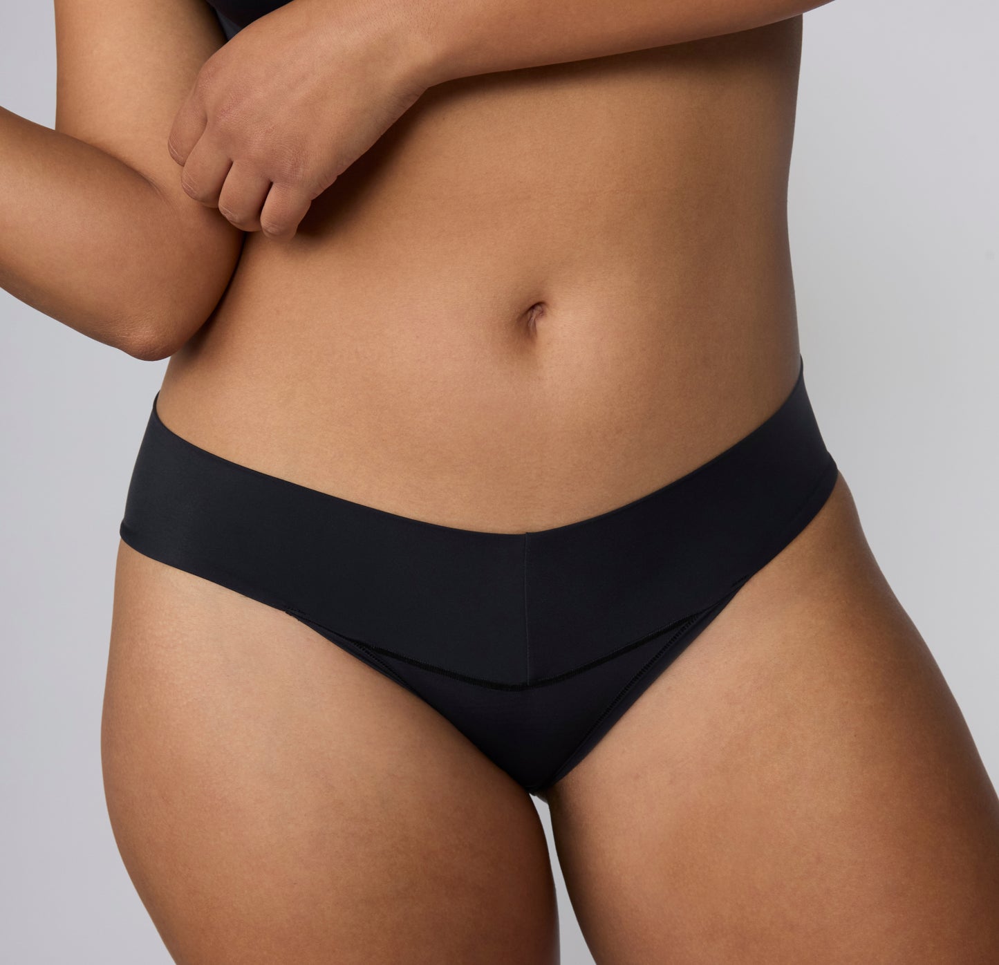 On a light gray studio backdrop, India wears the Bliss Low Rise Bikini in Black, shown in a close-up front view to highlight the smooth waistband, low-rise fit, and soft microfiber fabric.
