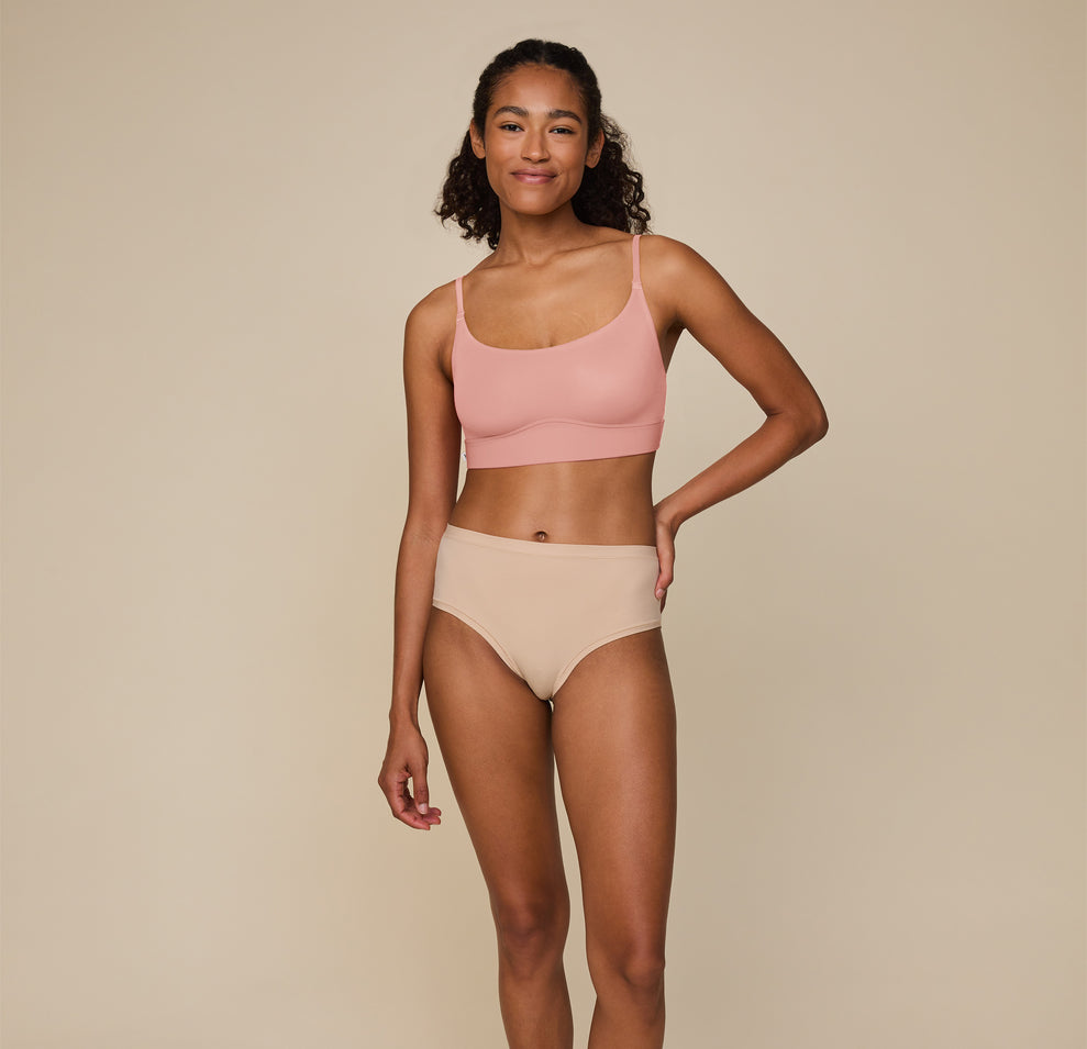 Full-body image of Zoe standing confidently, hand on her hip, in the Bliss Scoop Bralette in Dusty Rose and Bliss Highwaist Brief in Beige, showcasing the soft, relaxed fit of the matching set.