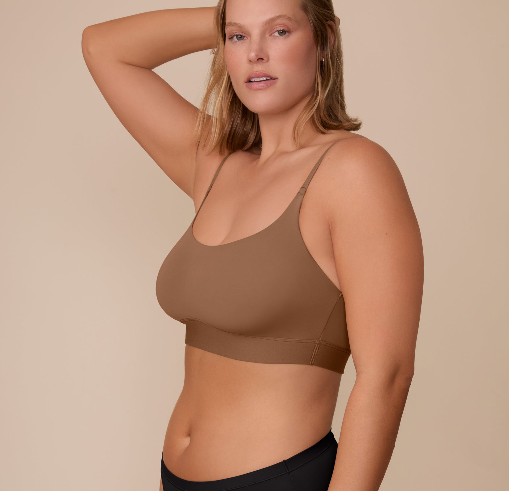 Side view of Sonnie wearing the Bliss Scoop Bralette in Brown and the Bliss Bikini in Black. One hand rests casually in her hair as she stands against a beige background.