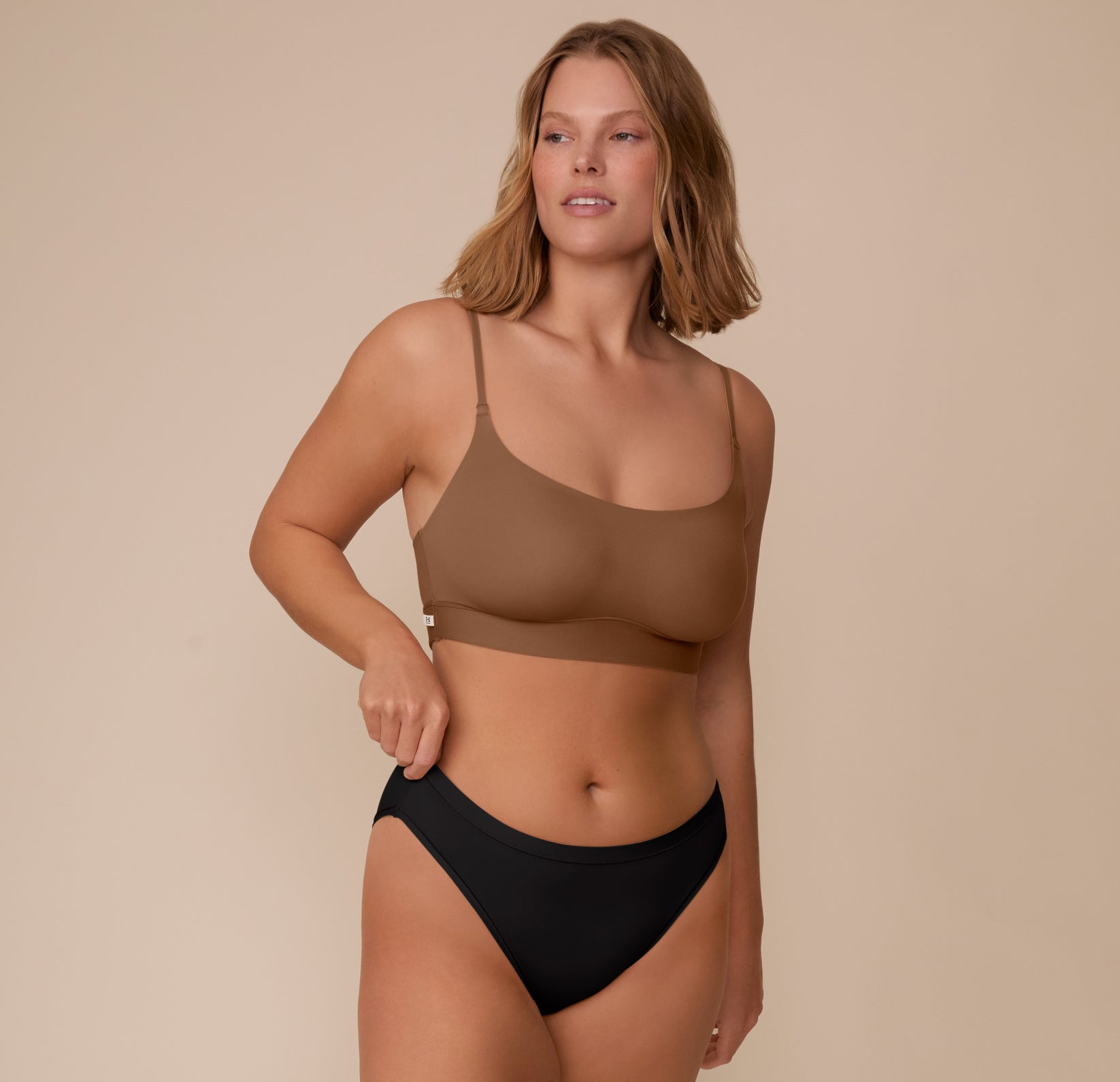 Sonnie wearing the Bliss Bralette in Brown and the Bliss Bikini in Black. One hand adjusts the waistband of the bikini brief. The wide underband and comfortable thin straps of the bralette are emphasized.