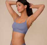 Marcela poses at a three quarter angle, casually running her hands through her hair. She wears the Bliss Scoop Bralette in Steel. The wide underband and thin straps are highlighted.