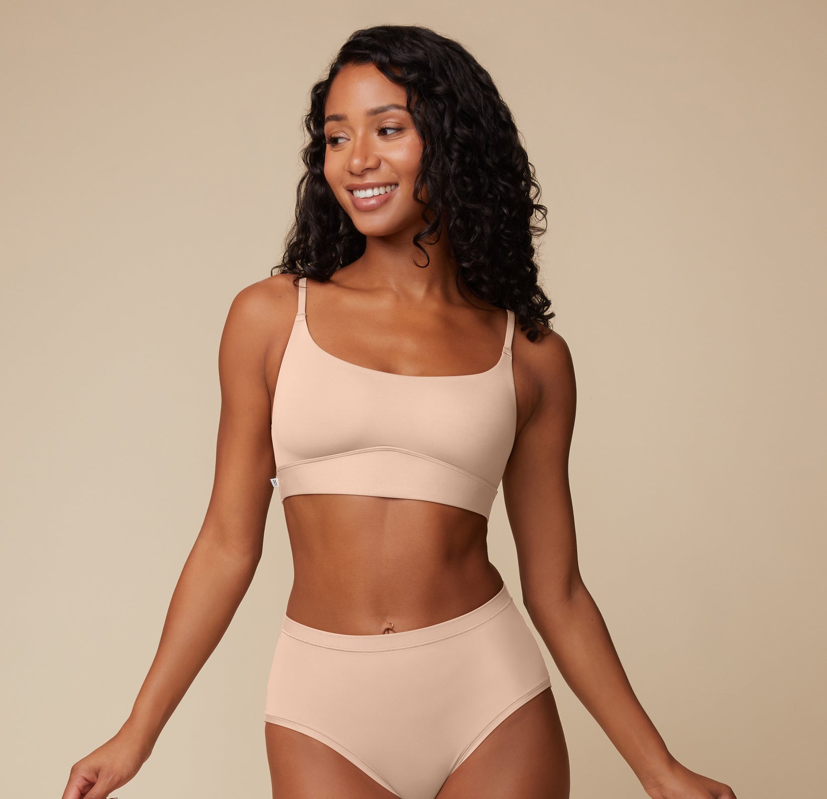Mid-length image of Ally smiling and wearing the Bliss Scoop Bralette and Bliss Highwaist Brief in Beige. The scoop neckline and full-coverage bottoms are clearly visible.