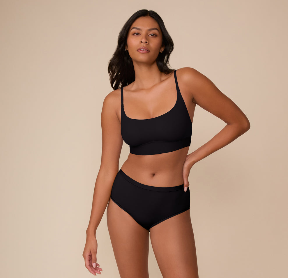 Full-body image of Victoria wearing the Bliss Scoop Bralette and Bliss Highwaist Brief in Black, standing confidently with one hand on hip.