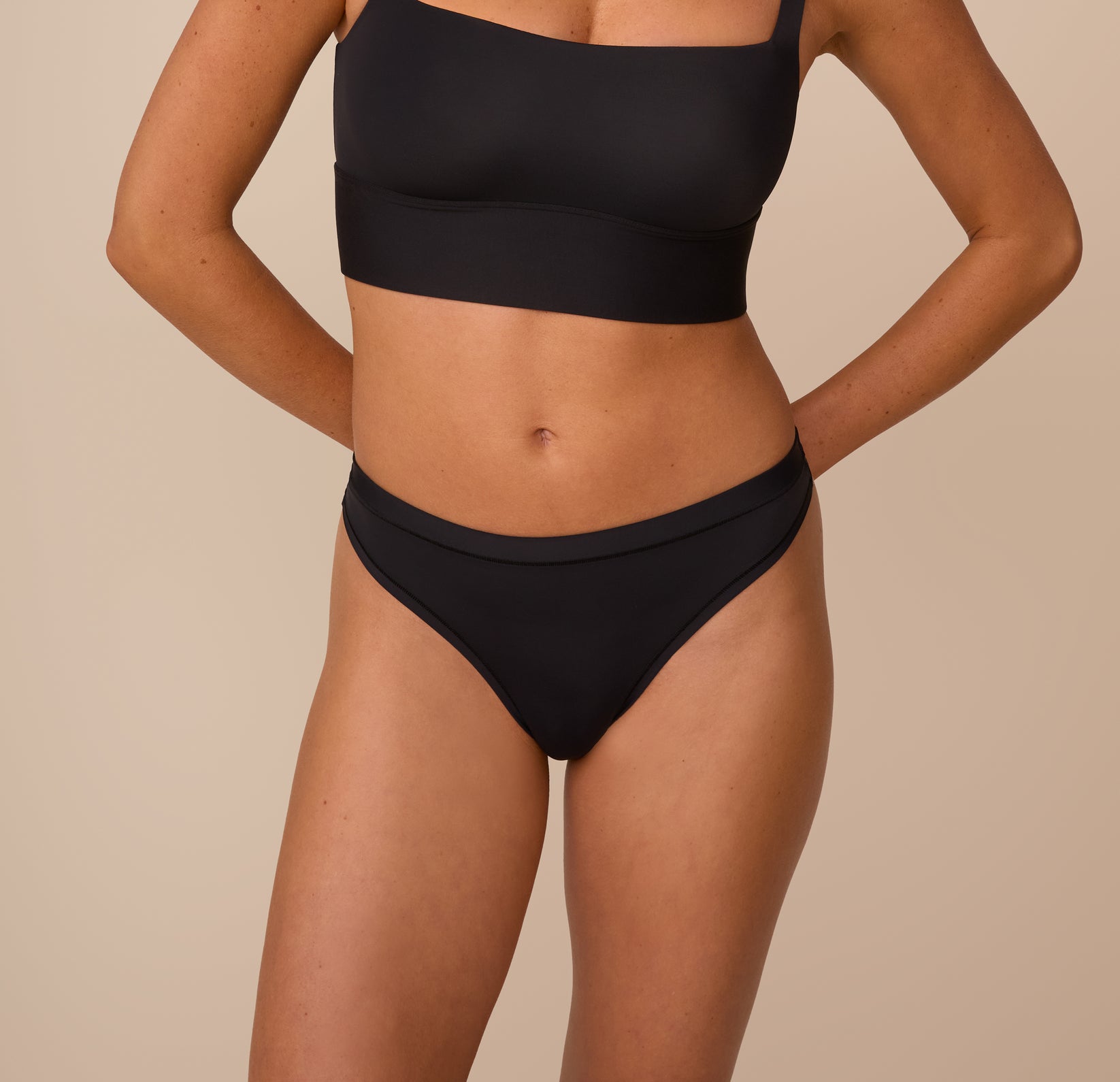 Freya wearing the Bliss Thong in Black, standing against a neutral beige background. The thong features a smooth, comfortable design with a low-rise fit and a soft waistband for a barely-there feel. sizing: Freya wears size Small