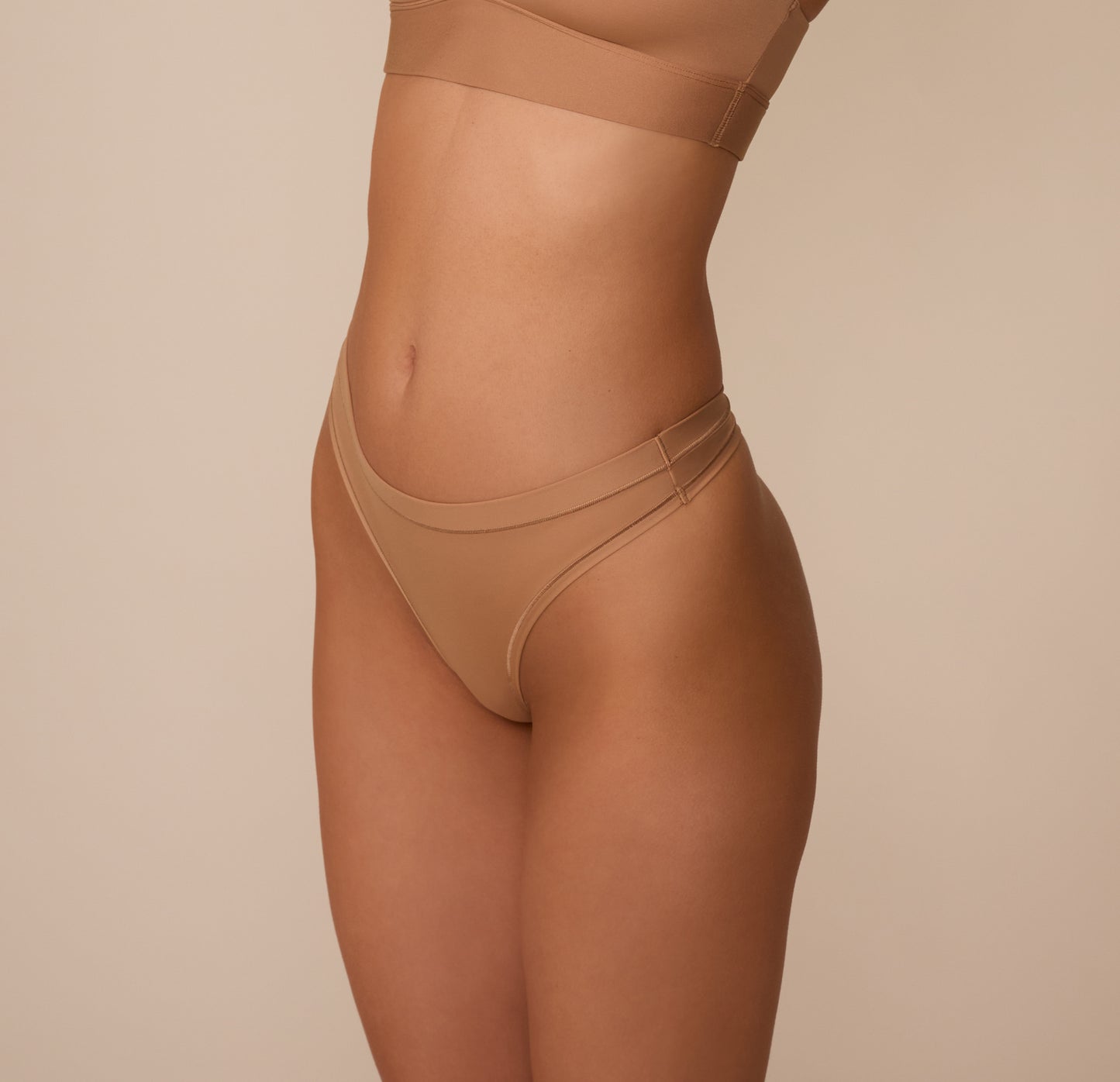 Three quarter view of Marcela wearing the Bliss Thong in Tan, showing the soft curve of the waistband and the stretch of the smooth, breathable fabric.