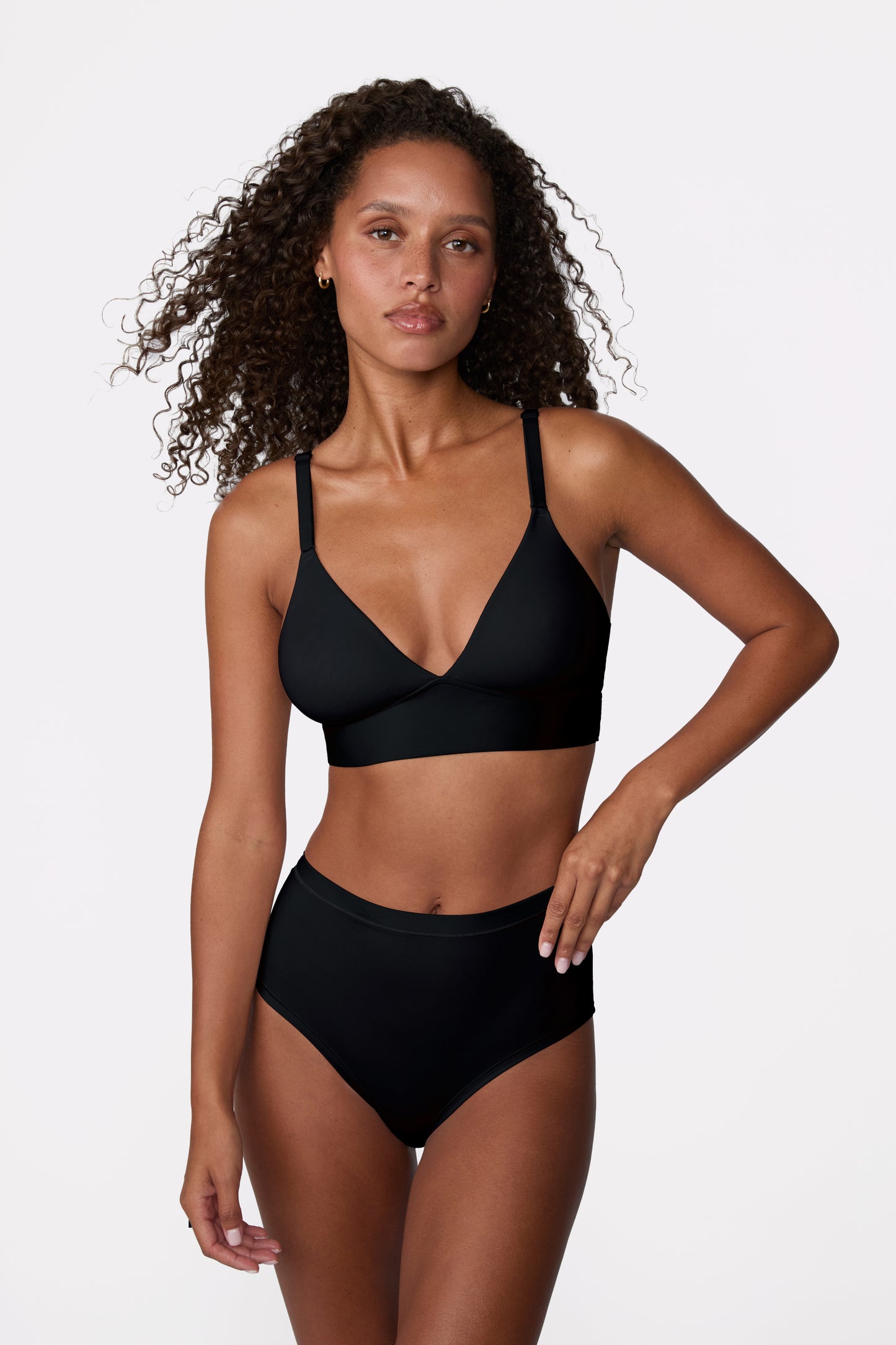 On a white studio backdrop, Courtney stands facing forward wearing the Bliss Triangle Bralette in Black with matching bottoms, showing the low-coverage silhouette, adjustable straps, and smooth microfiber fabric designed for everyday comfort.