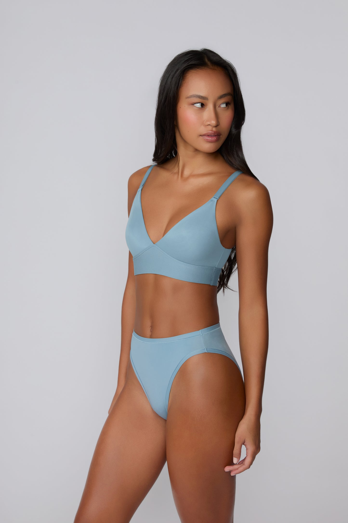 Three-quarter front view on a light gray studio backdrop of Amber wearing the Bliss Triangle Bralette in Poolside with the coordinating bikini brief, showing the deep V neckline, wireless pullover design, and adjustable straps.