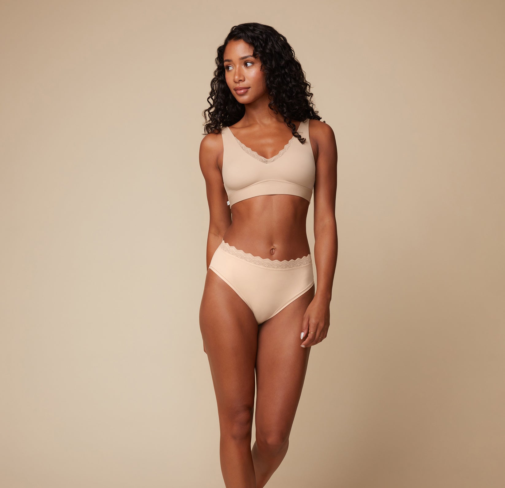 Model Ally wearing the Bliss Lace Bralette in Beige paired with the Bliss Lace Highwaist Brief in Beige. She stands facing forward, showcasing the comfortable, supportive bralette and highwaist brief with full coverage.