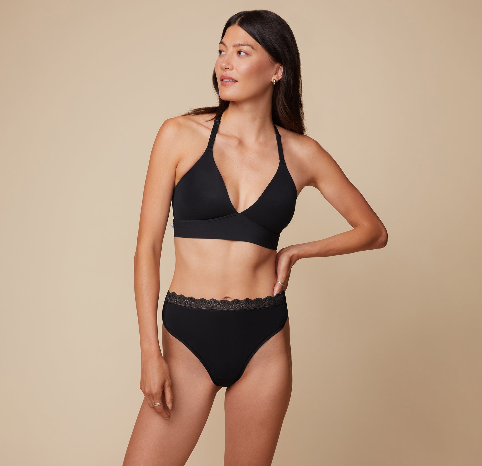 Anna posing in the Bliss Lace Halterneck Bralette and matching Bliss Highwaist Lace Thong in Black. The image highlights the bralette’s halterneck design and the thong’s high-waist fit, with Anna gazing to the side.