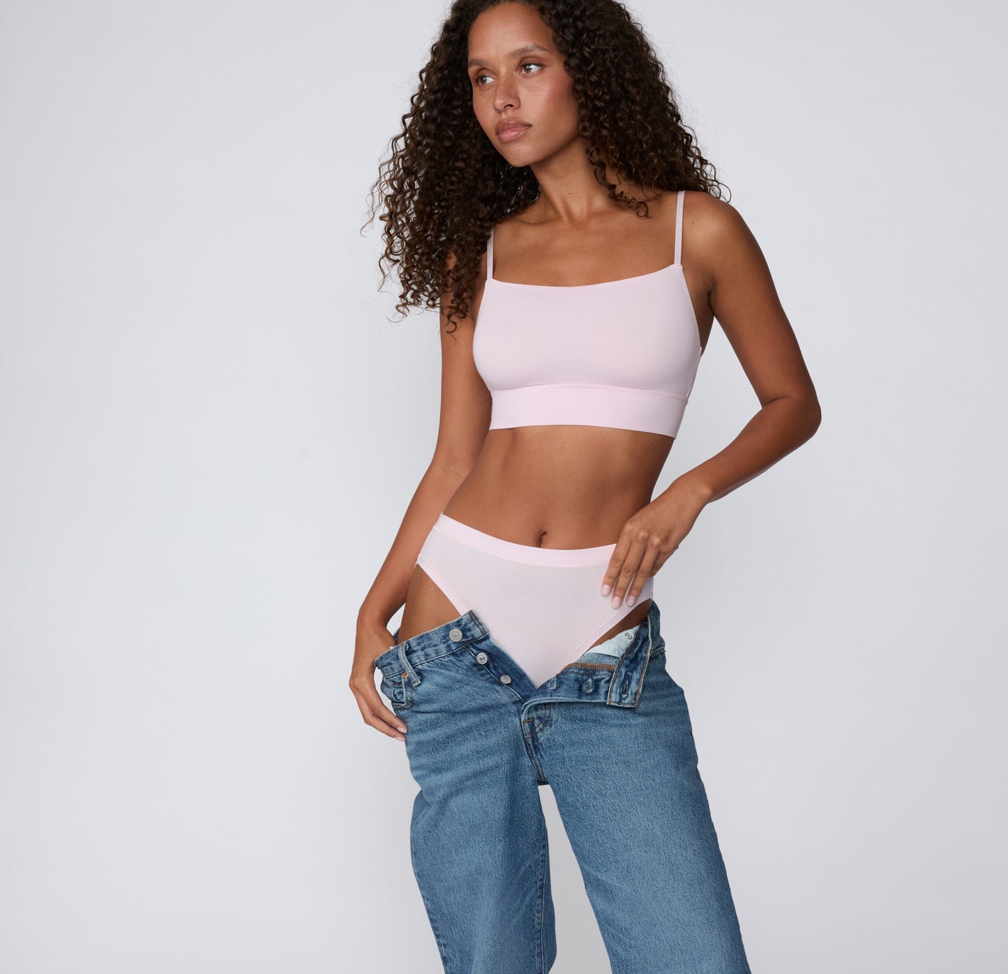 On a clean studio backdrop, a full-body view shows a model wearing the Cloud Cotton French Cut Brief in Primrose with the matching Cloud Cotton Thin Strap Bralette, styled with low-rise denim.