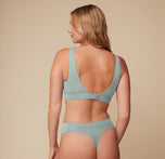 Megan is shown from the back, wearing the Cloud Cotton Scoop Bralette in seaglass, which features an adjustable hook closure for a customized fit. She pairs it with matching high-waisted thong, against a beige background.