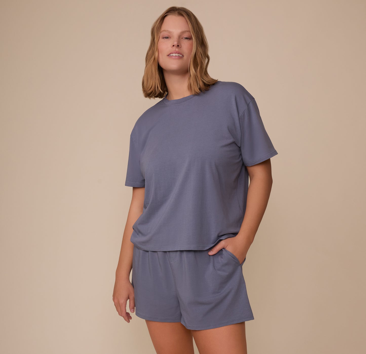 Full body image of Sonnie wearing the Cloud Cotton Sleep Short and matching T-Shirt in Slate. She smiles softly with one hand in her pocket, showing off the easygoing, cozy sleepwear set.